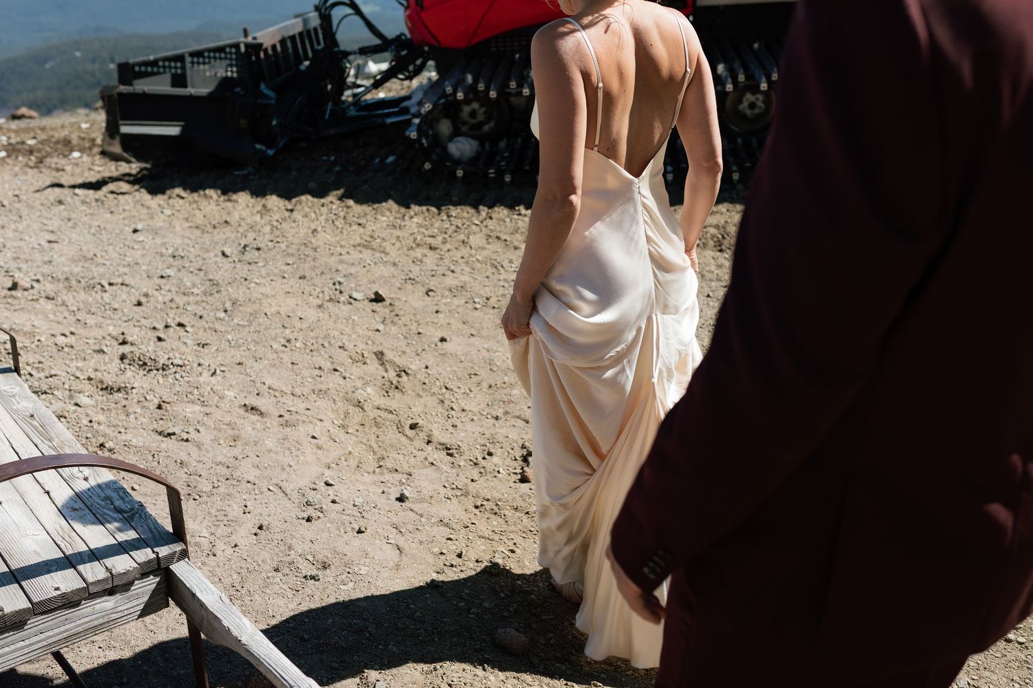 Bride in elegant backless satin gown walks with groom at mountain wedding.