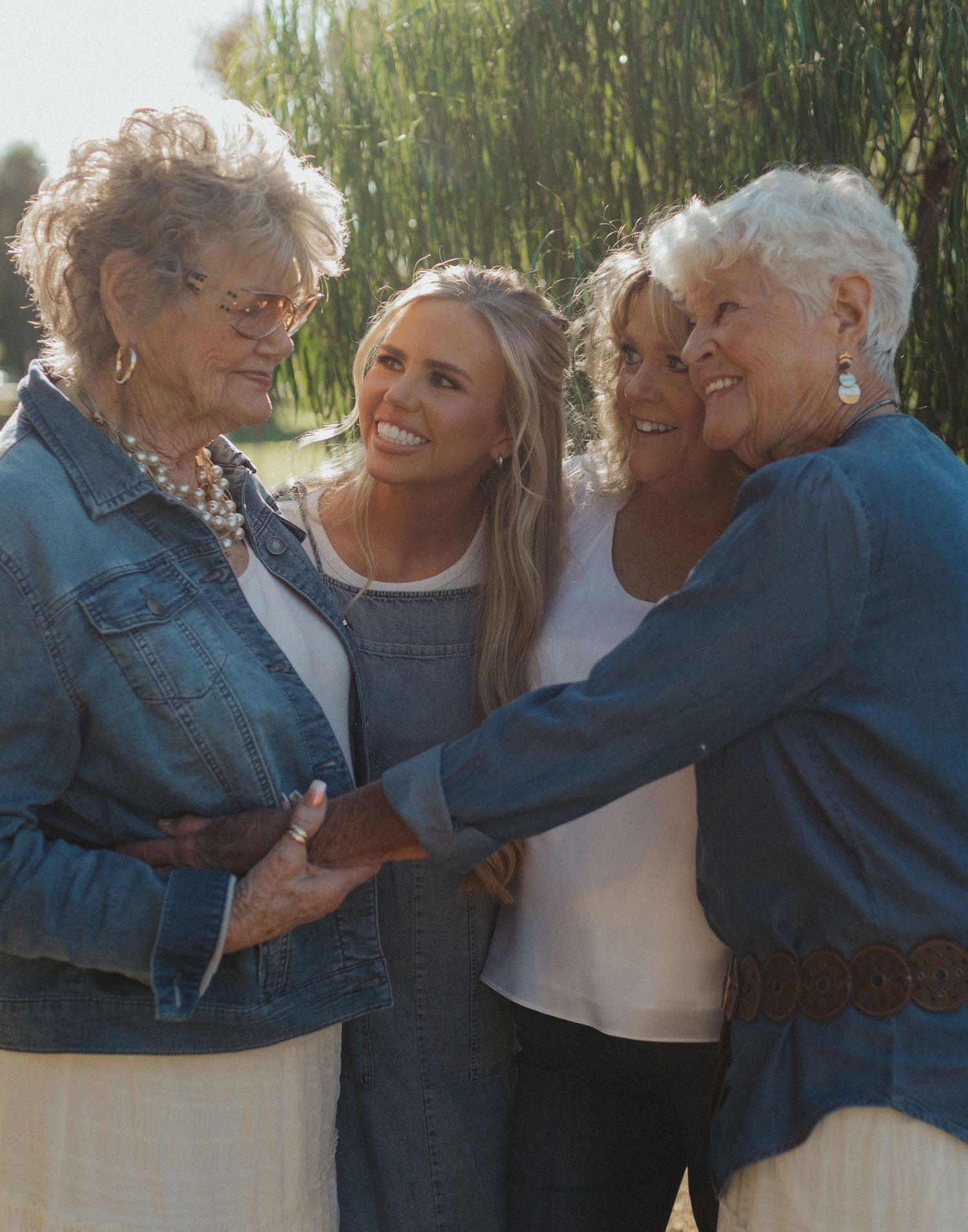 Four women of different generations laugh and embrace together outdoors in warm sunlight.