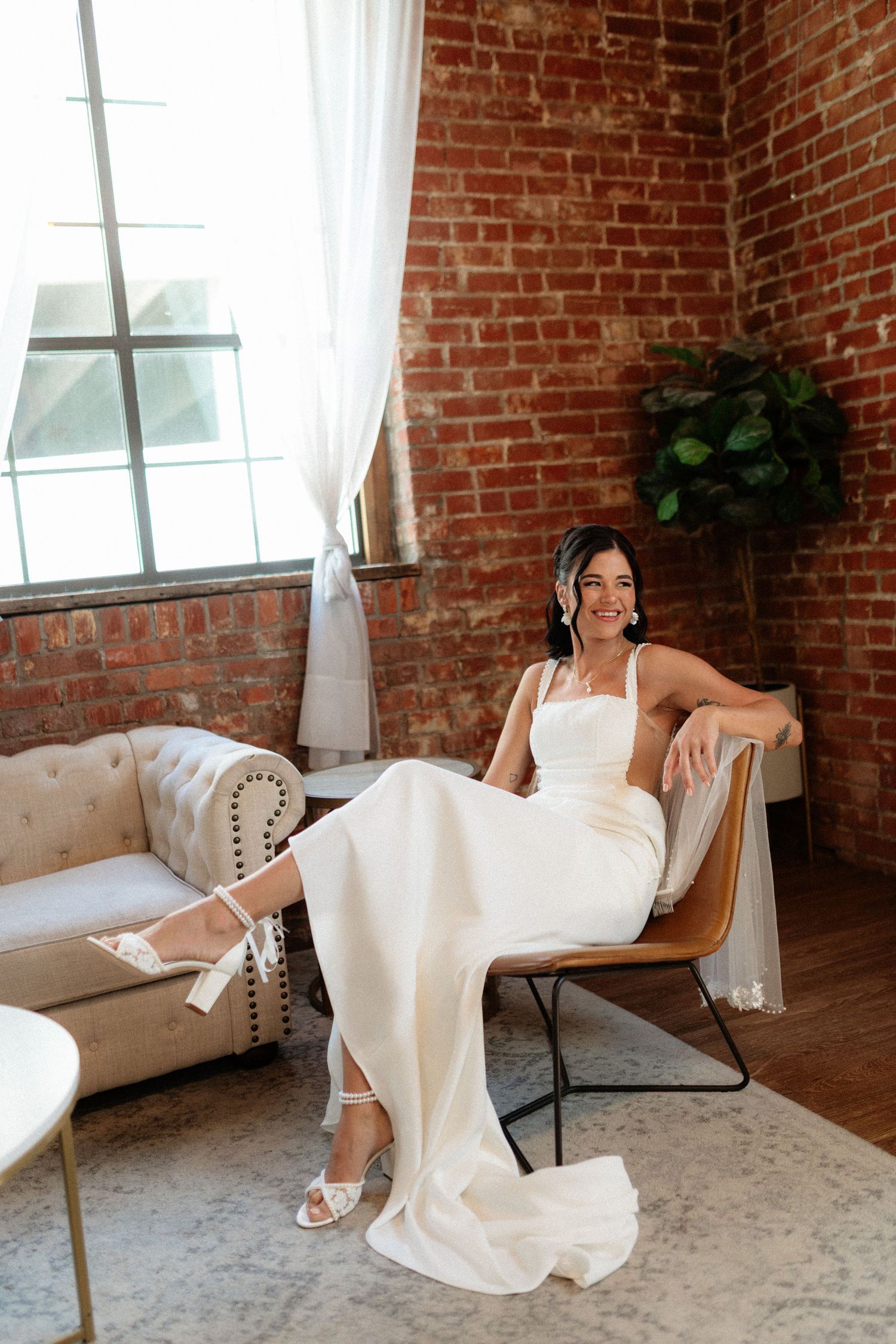 Smiling bride in sleek white gown and strappy heels poses seated in a chair in a brick-walled loft space.