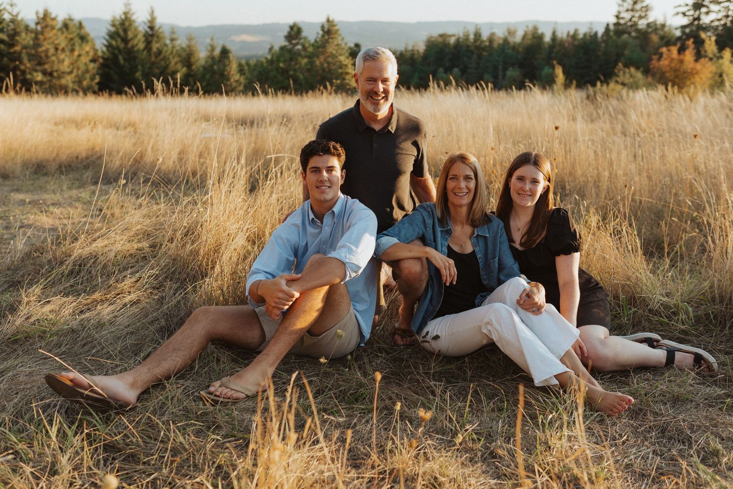 Family of four posing together in a sunny dry grass field with evergreen trees behind.