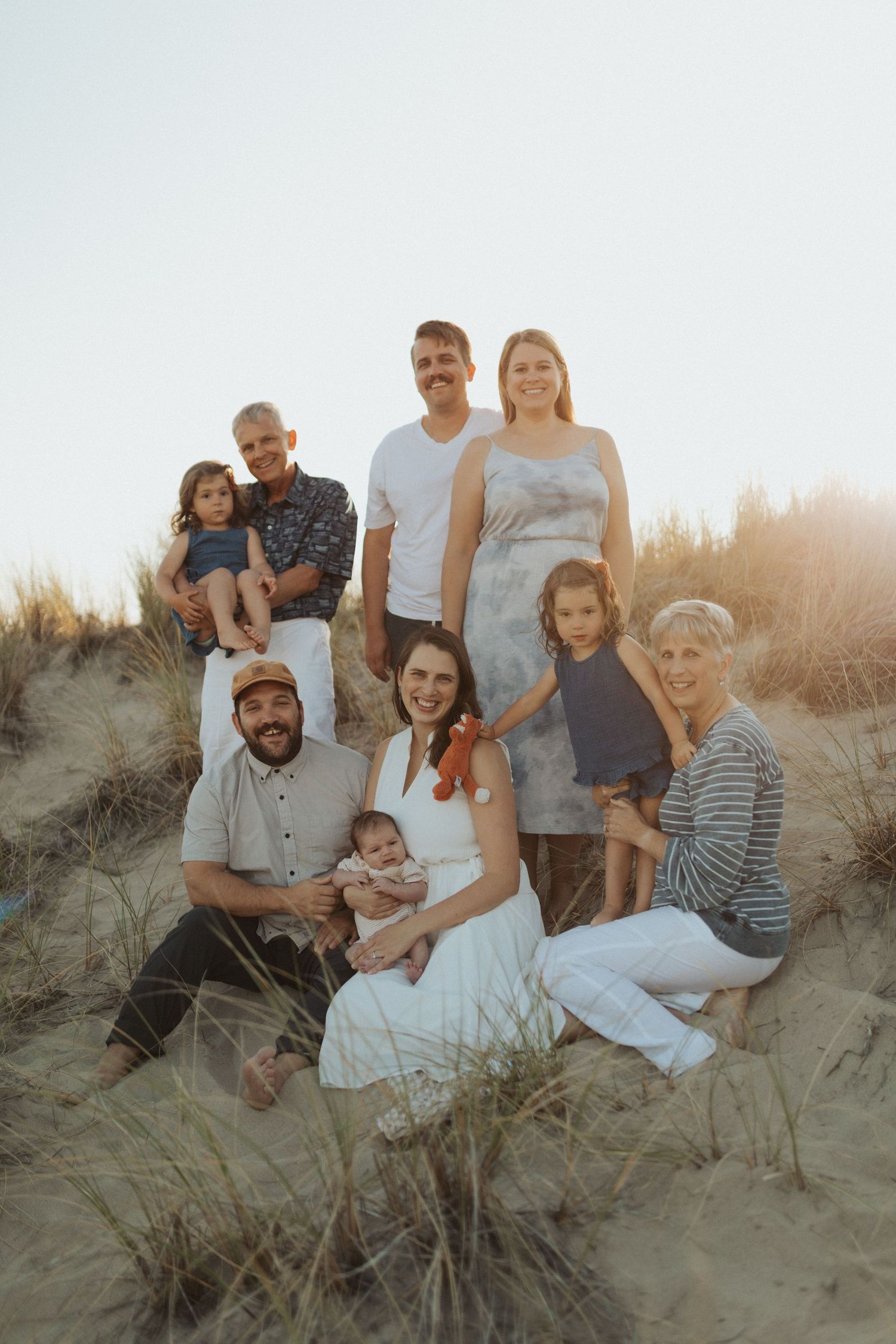Extended family portrait on sandy beach dunes at golden hour sunset with eight members posing together.