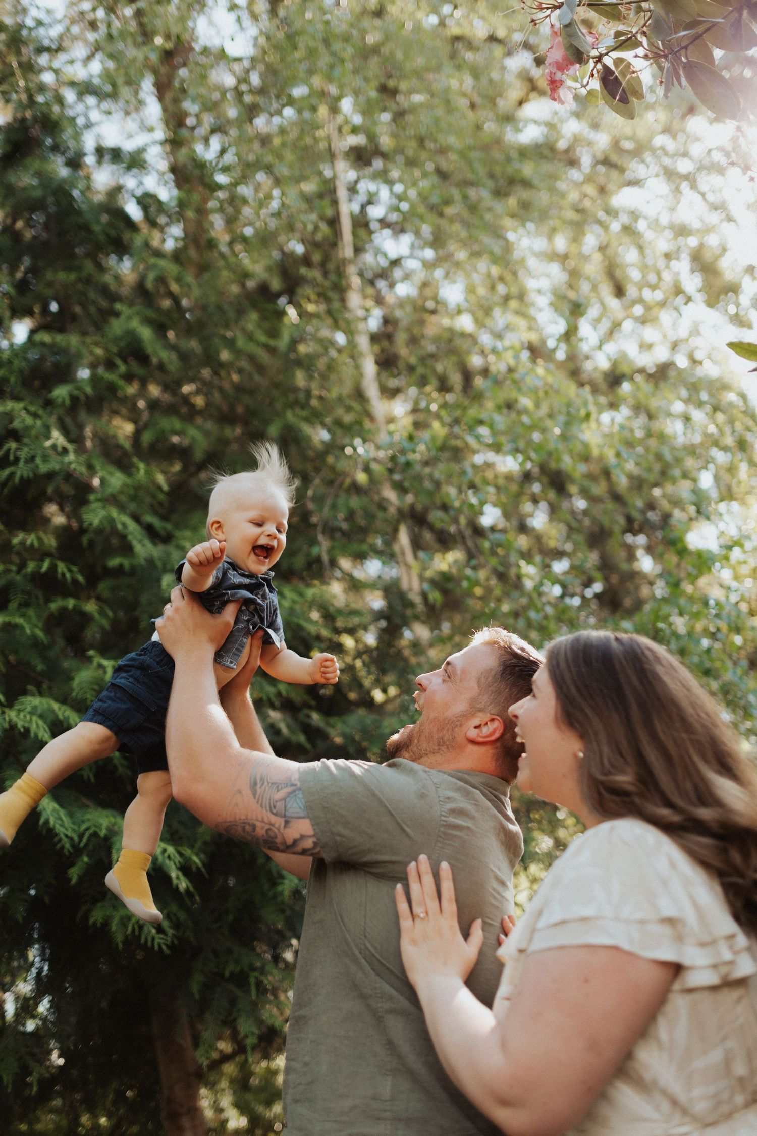Joyful parents lift their laughing baby into the air outdoors among green trees and sunlight.