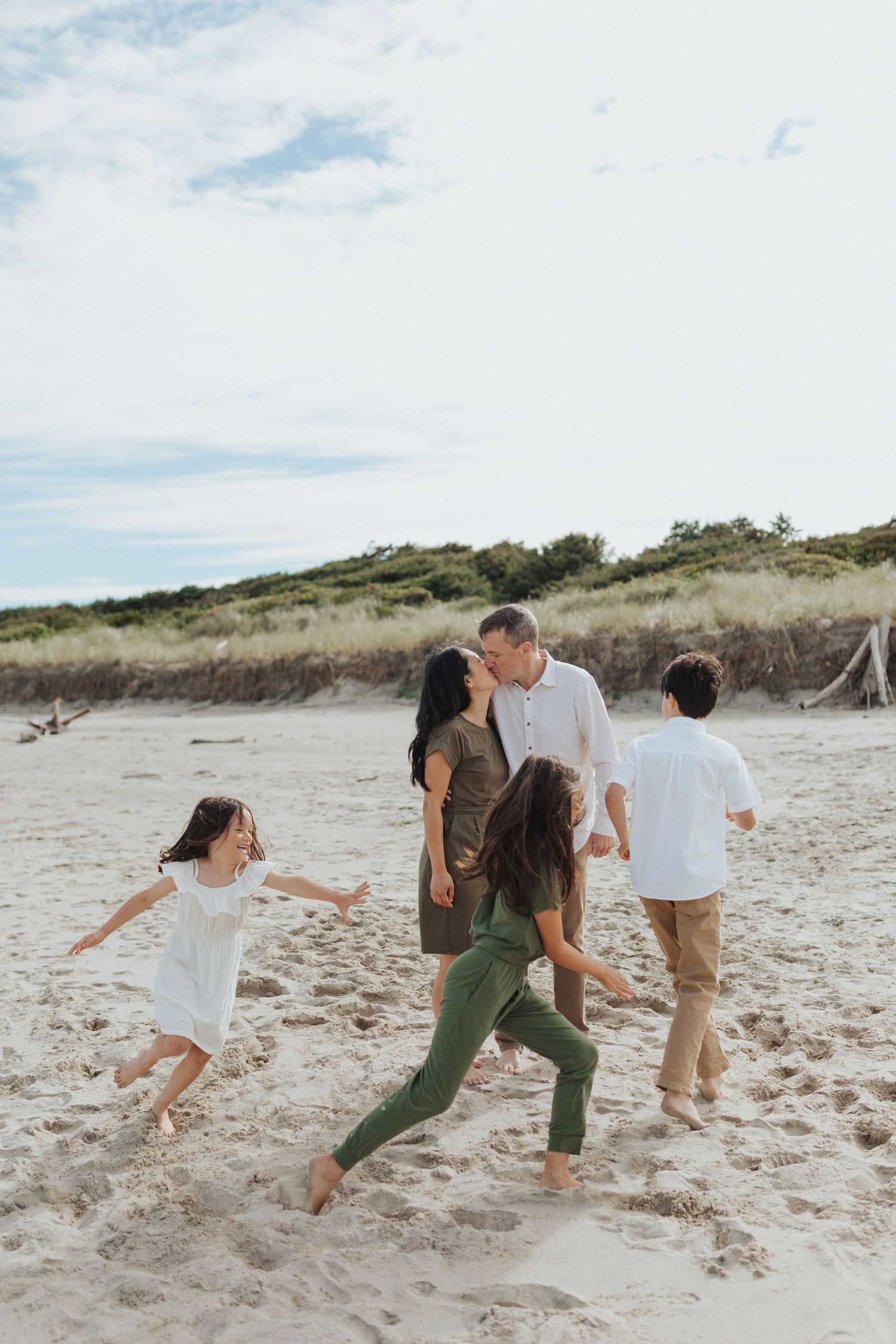 Family of five on a sandy beach, parents kissing while three children run and play around them.