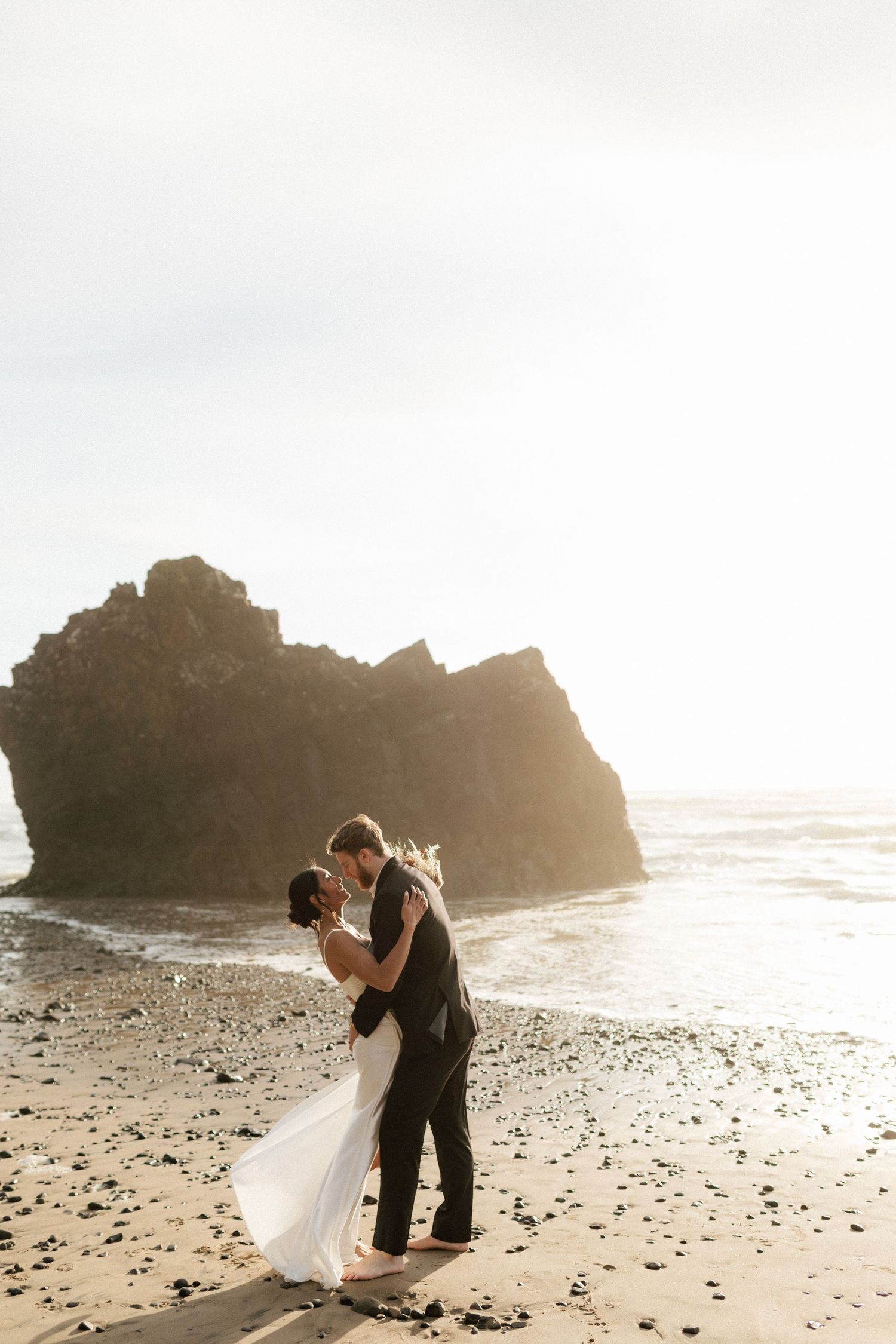 Bride and groom share a romantic kiss on a scenic beach with dramatic rock formations and ocean waves in the background.