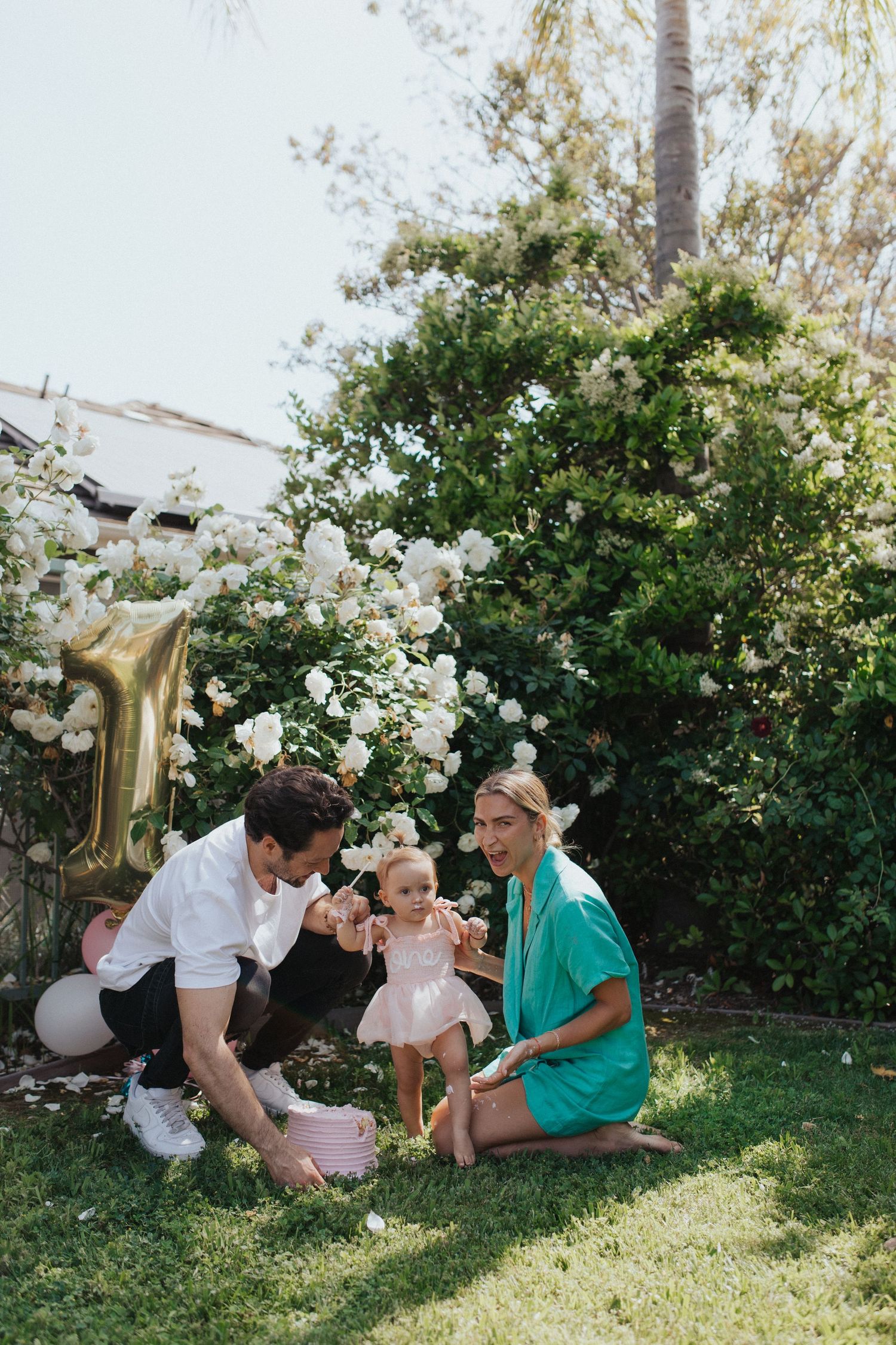 Young family with baby girl celebrate first birthday outdoors near white rose bush and gold balloon.