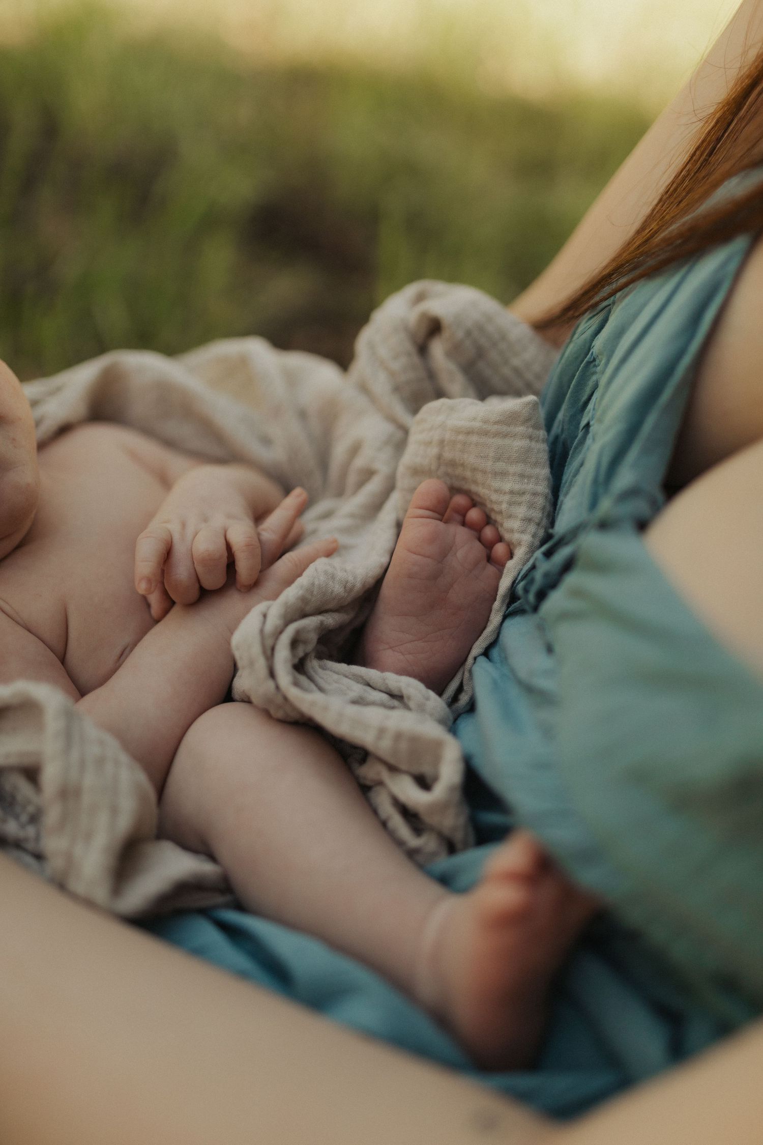 Close-up of newborn baby feet and hands wrapped in soft muslin blanket on mother's lap outdoors.