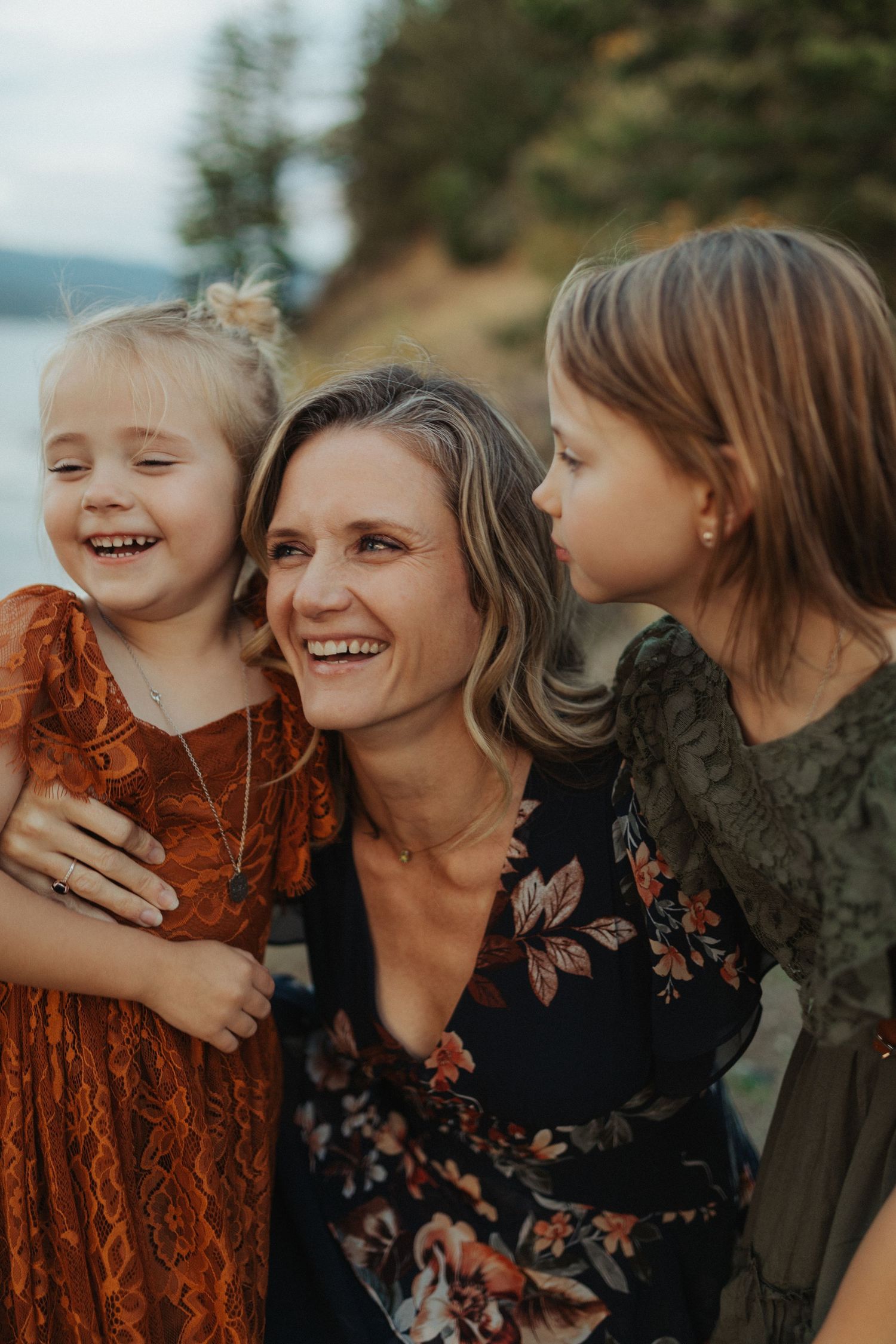 A joyful mother laughs as her two daughters cuddle close to her cheeks near a lakeside setting.