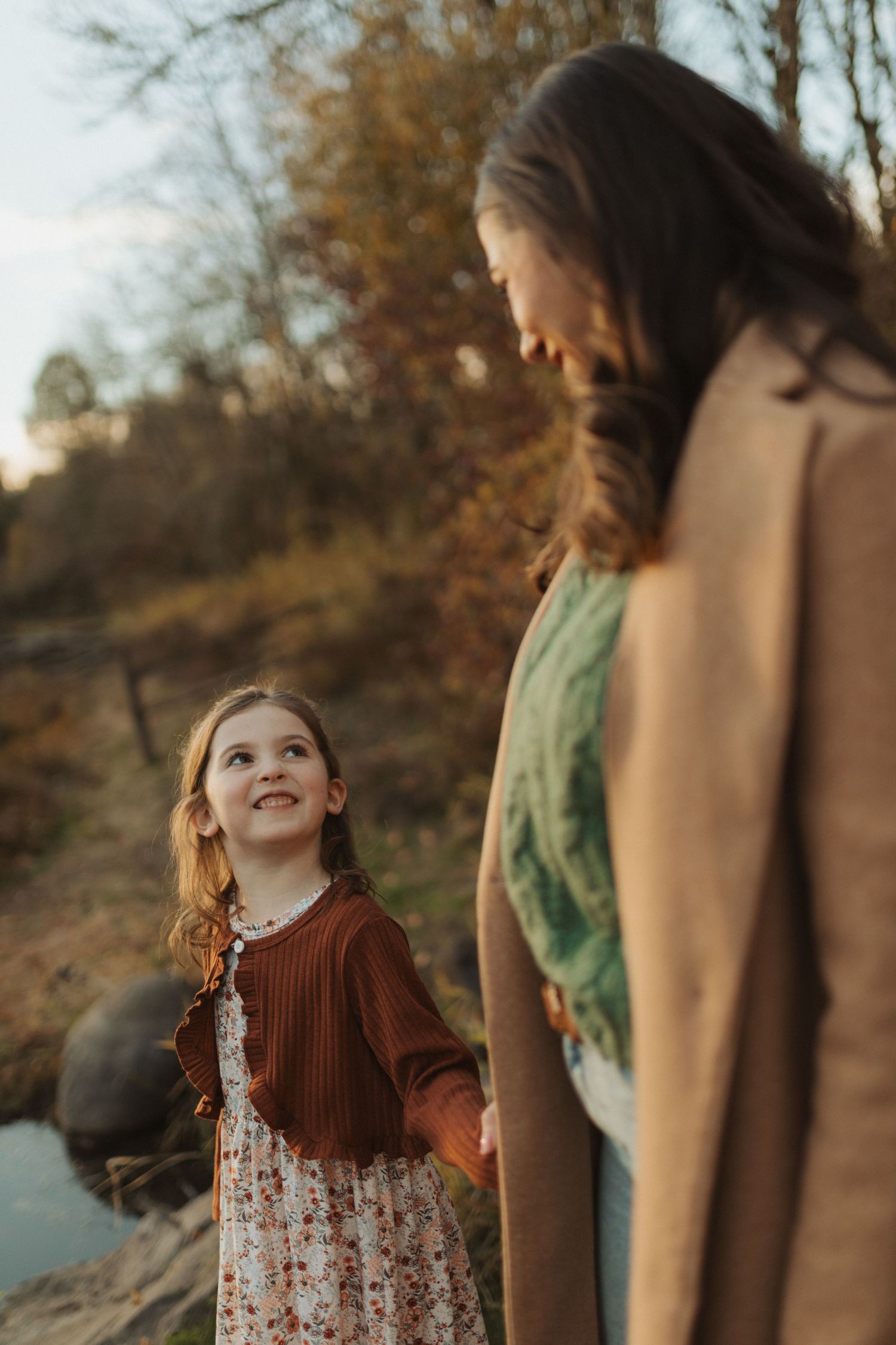 A young girl in a floral dress and rust cardigan smiles up at her mother during an autumn outdoor photo session.