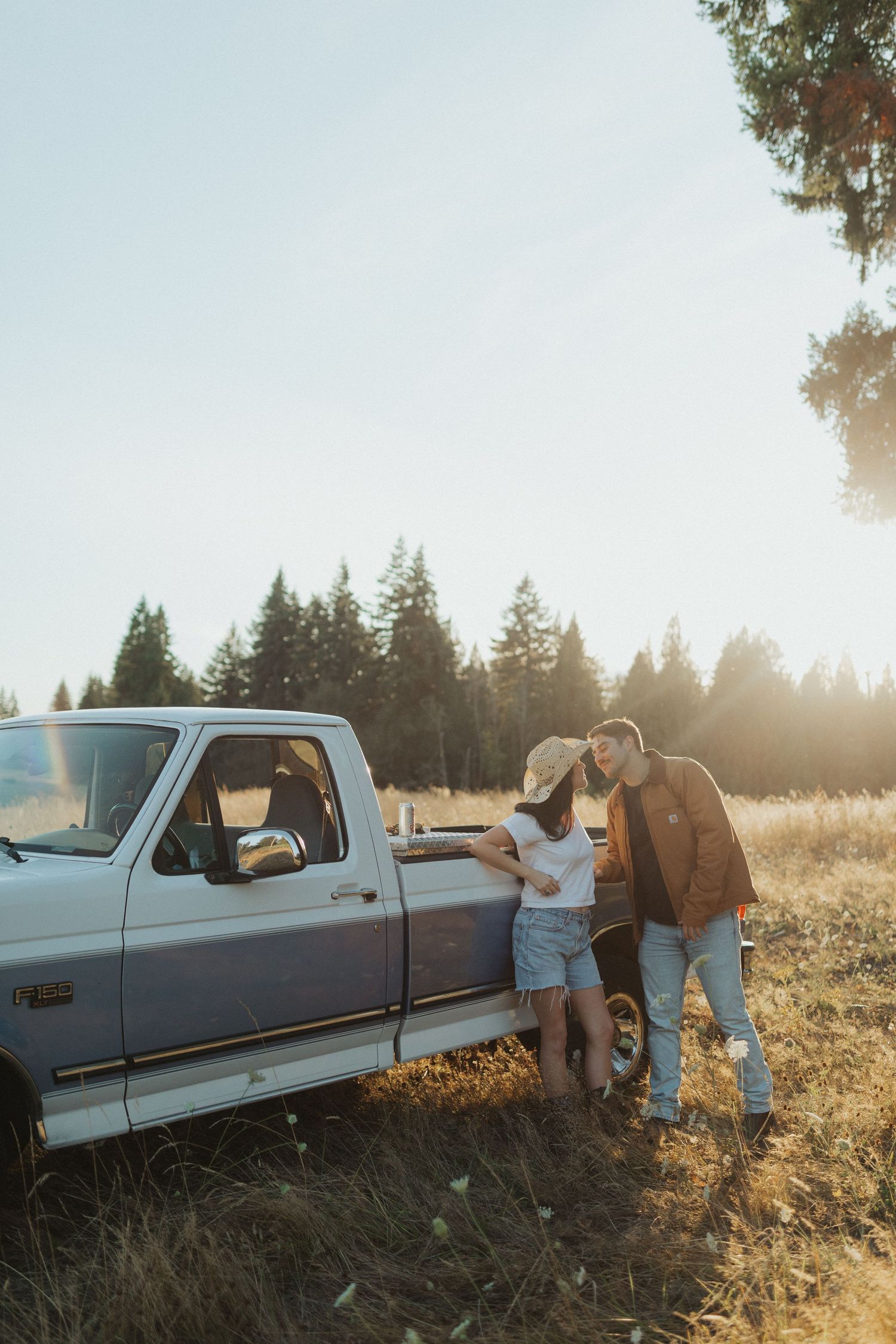 Couple sharing a romantic moment leaning against a vintage white pickup truck in a sunlit field.