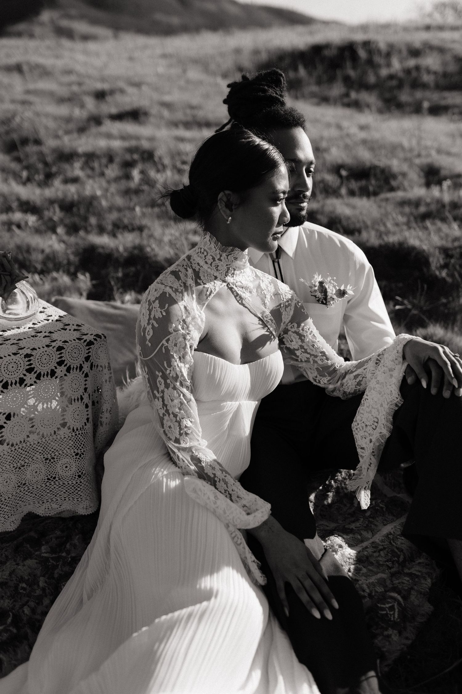 Black and white portrait of bride in lace wedding gown and groom sitting together on a grassy hillside.