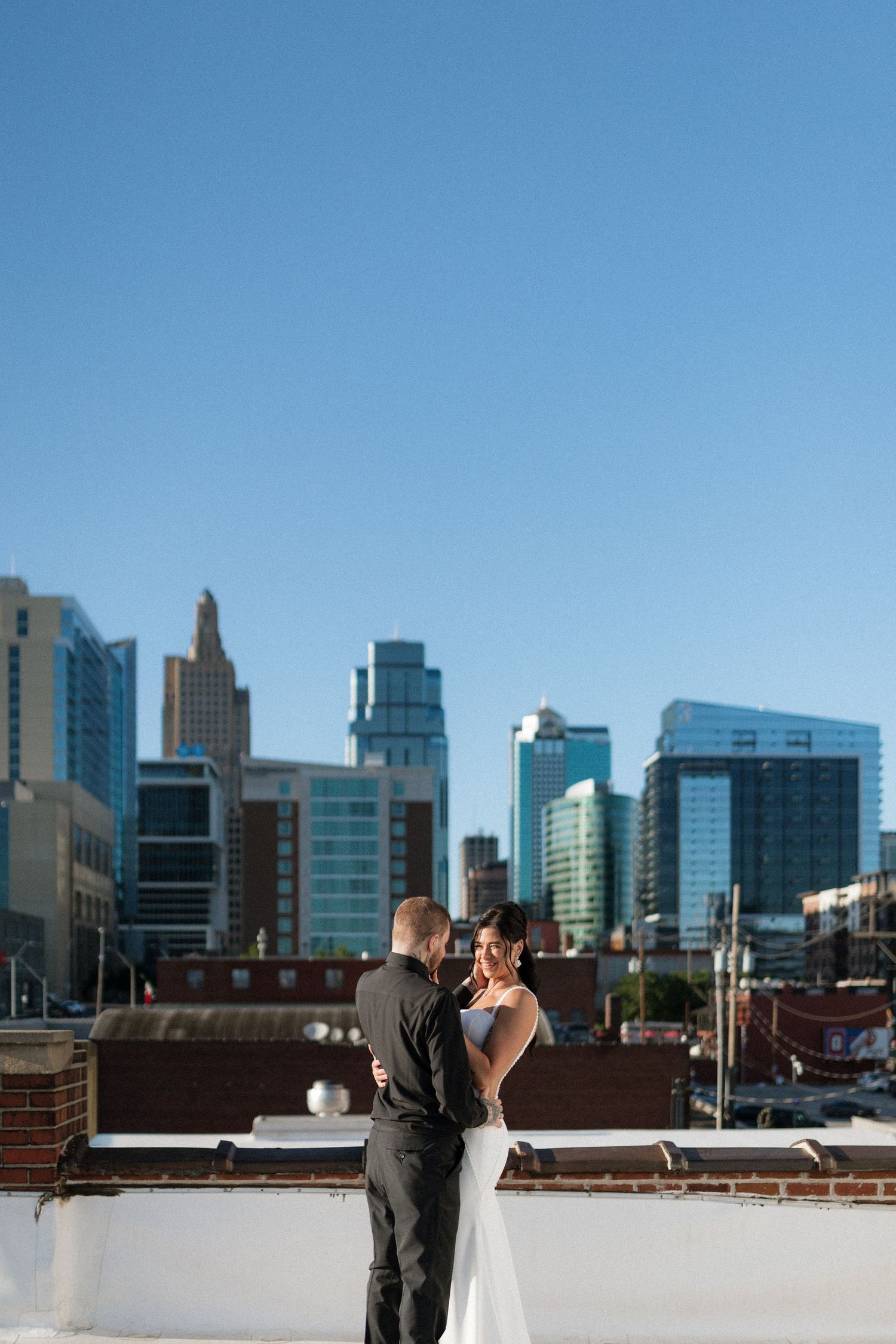 A bride and groom embrace on a rooftop with a stunning urban city skyline visible in the background.