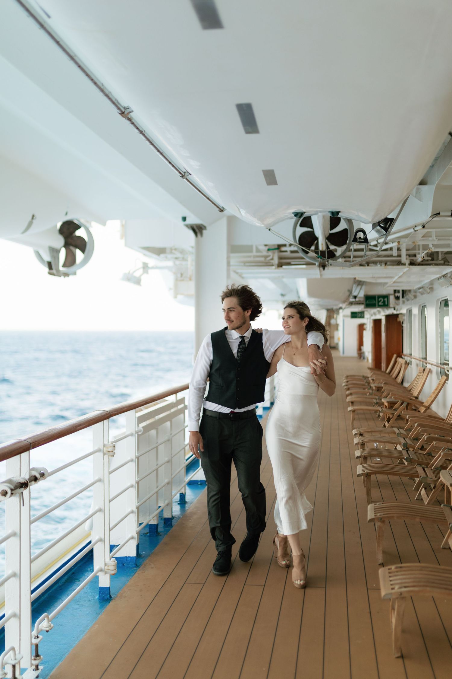 Bride and groom walk arm in arm along a cruise ship deck overlooking the ocean.