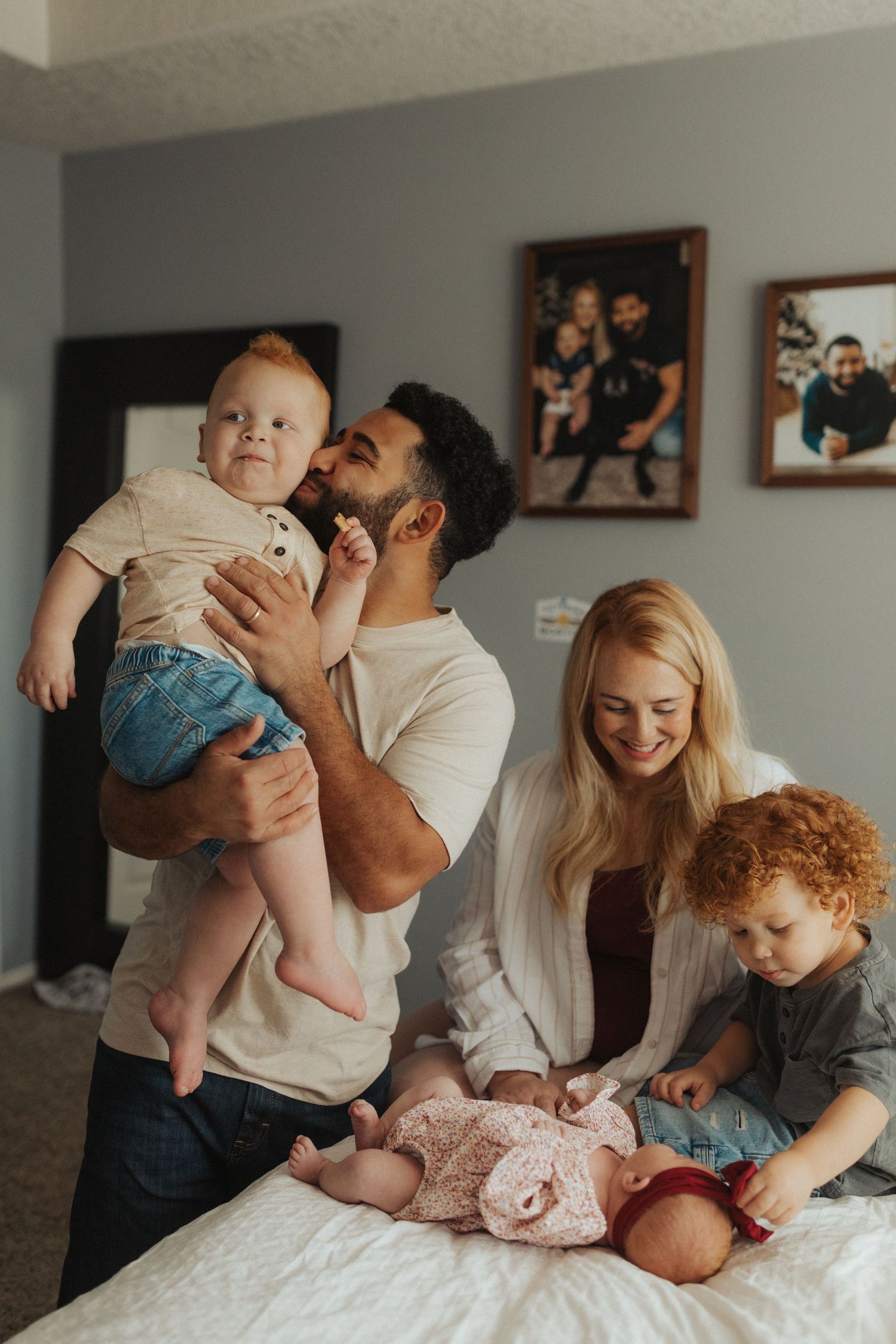A father kisses a baby he holds up while the mother and curly-haired toddler smile on a bed.