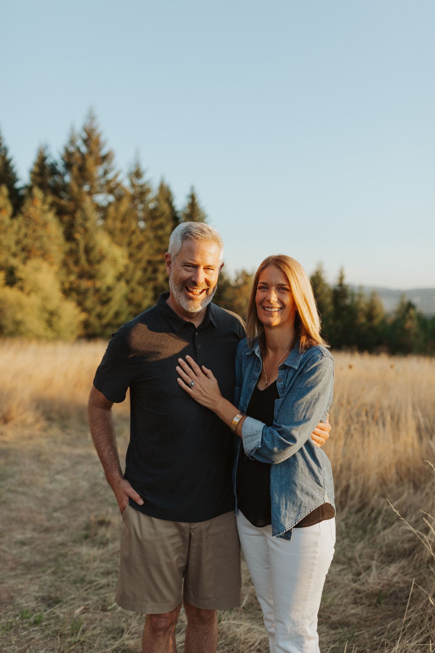 Smiling couple embracing in a golden field with evergreen trees at sunset.