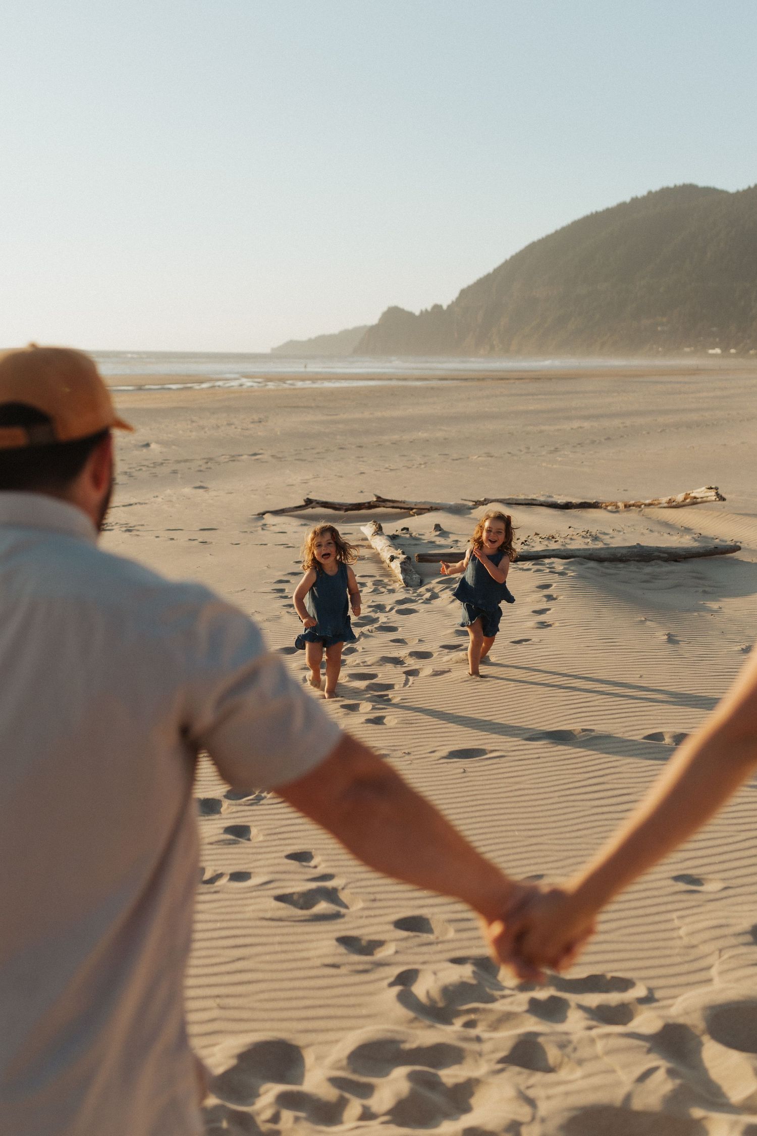 Father holding hands with partner while two young girls run toward him on a sunny beach.
