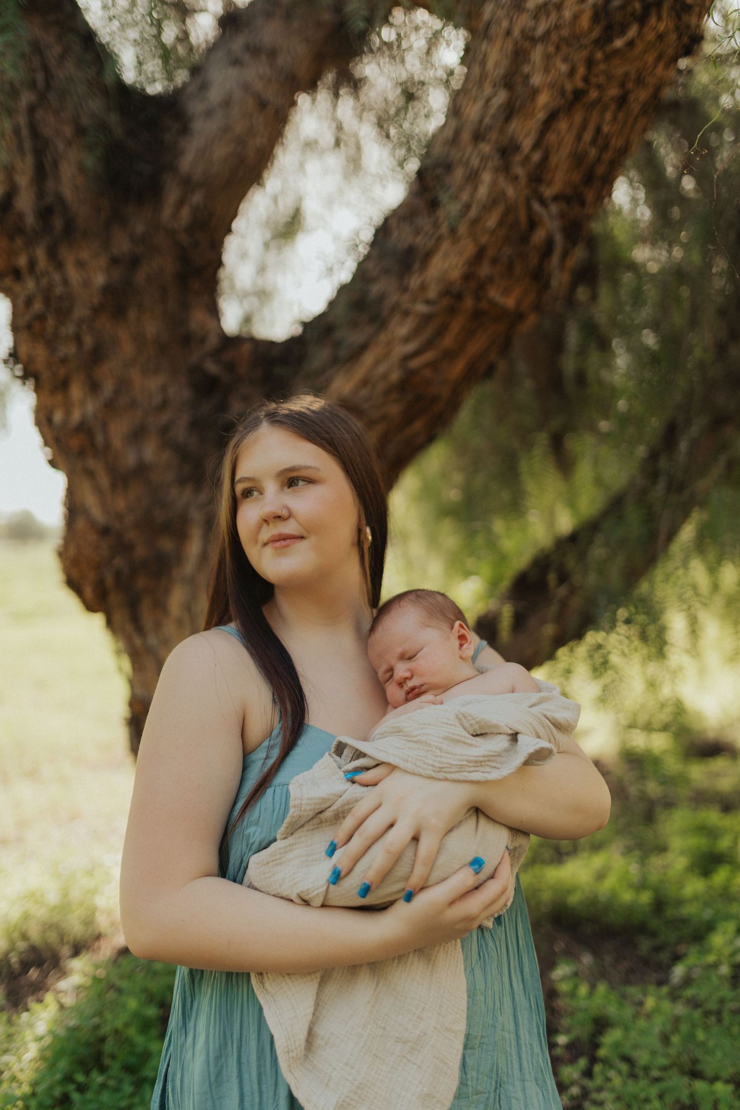New mother in blue dress cradling sleeping newborn wrapped in cream blanket outdoors near tree.