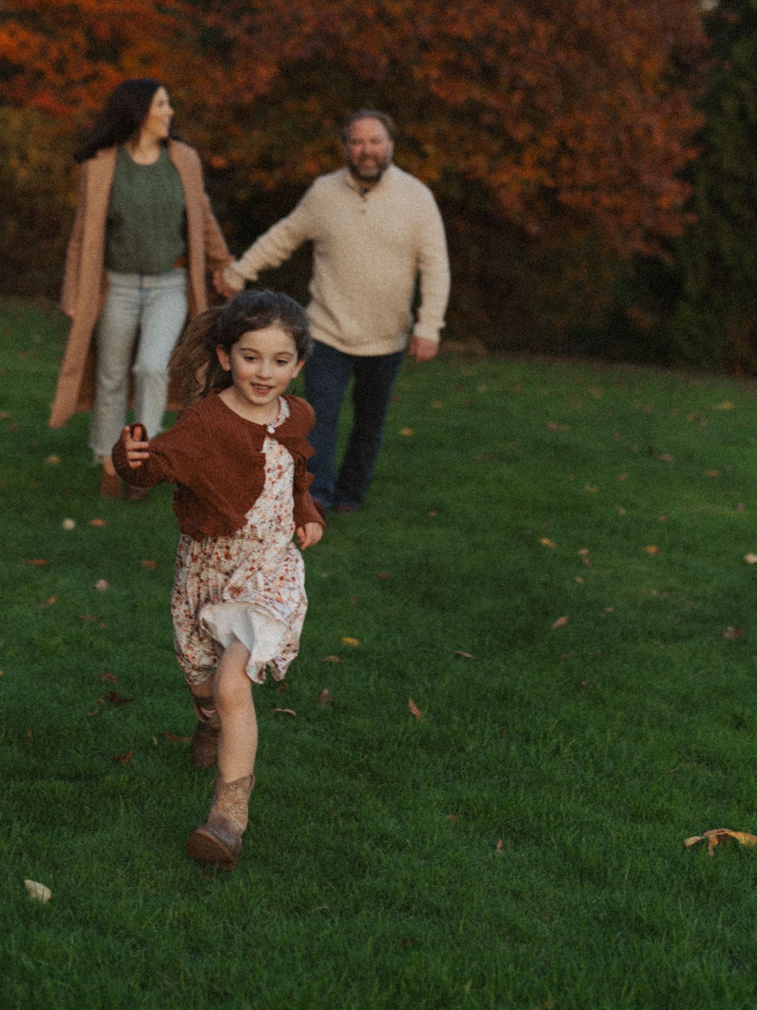 A young girl in a floral dress runs across a green lawn while her parents hold hands in the background.