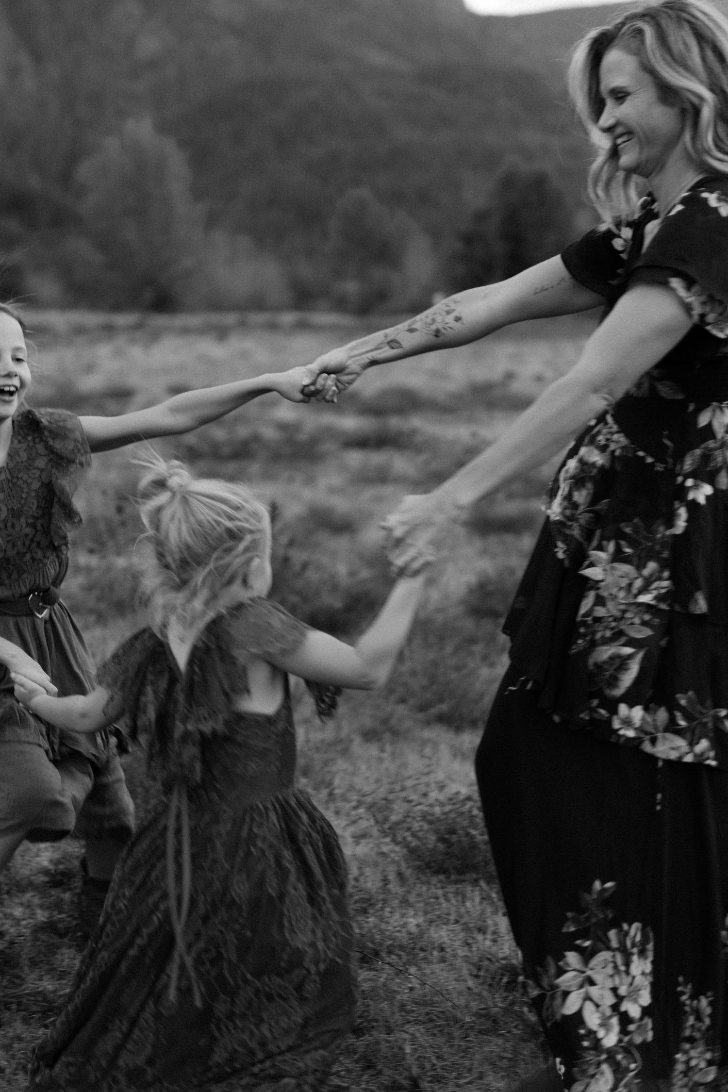 Black and white photo of a laughing mother spinning her two daughters by their hands in a field.