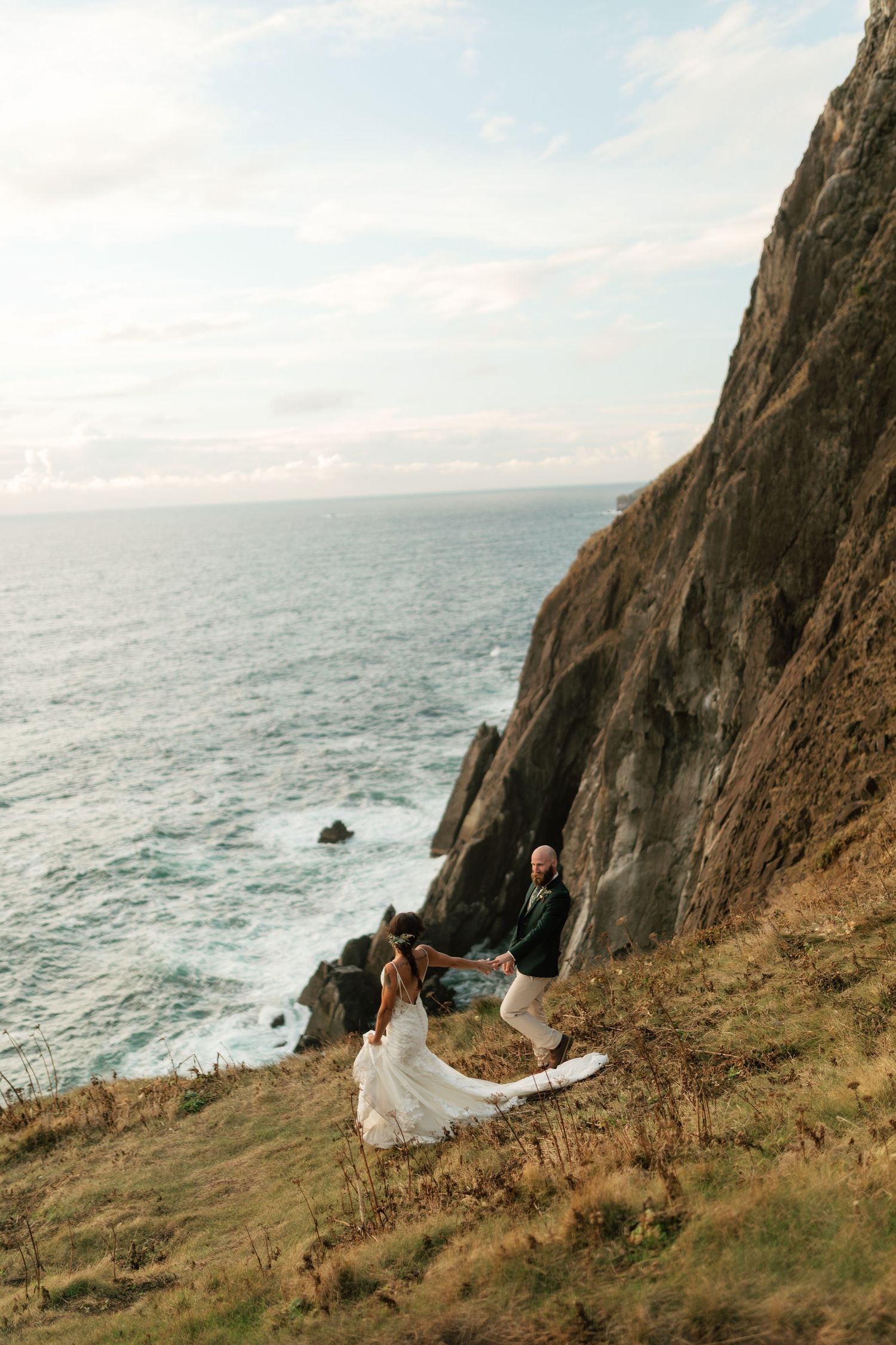 Bride and groom standing on dramatic coastal cliff edge with crashing ocean waves and rocky cliffs below.