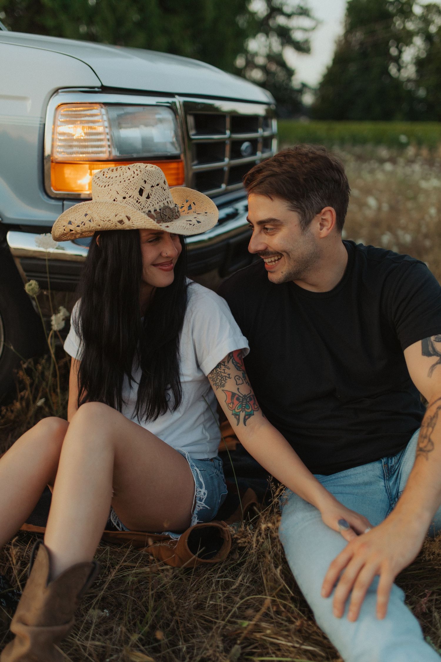 A happy couple sits together in a field in front of a vintage Ford truck at dusk.
