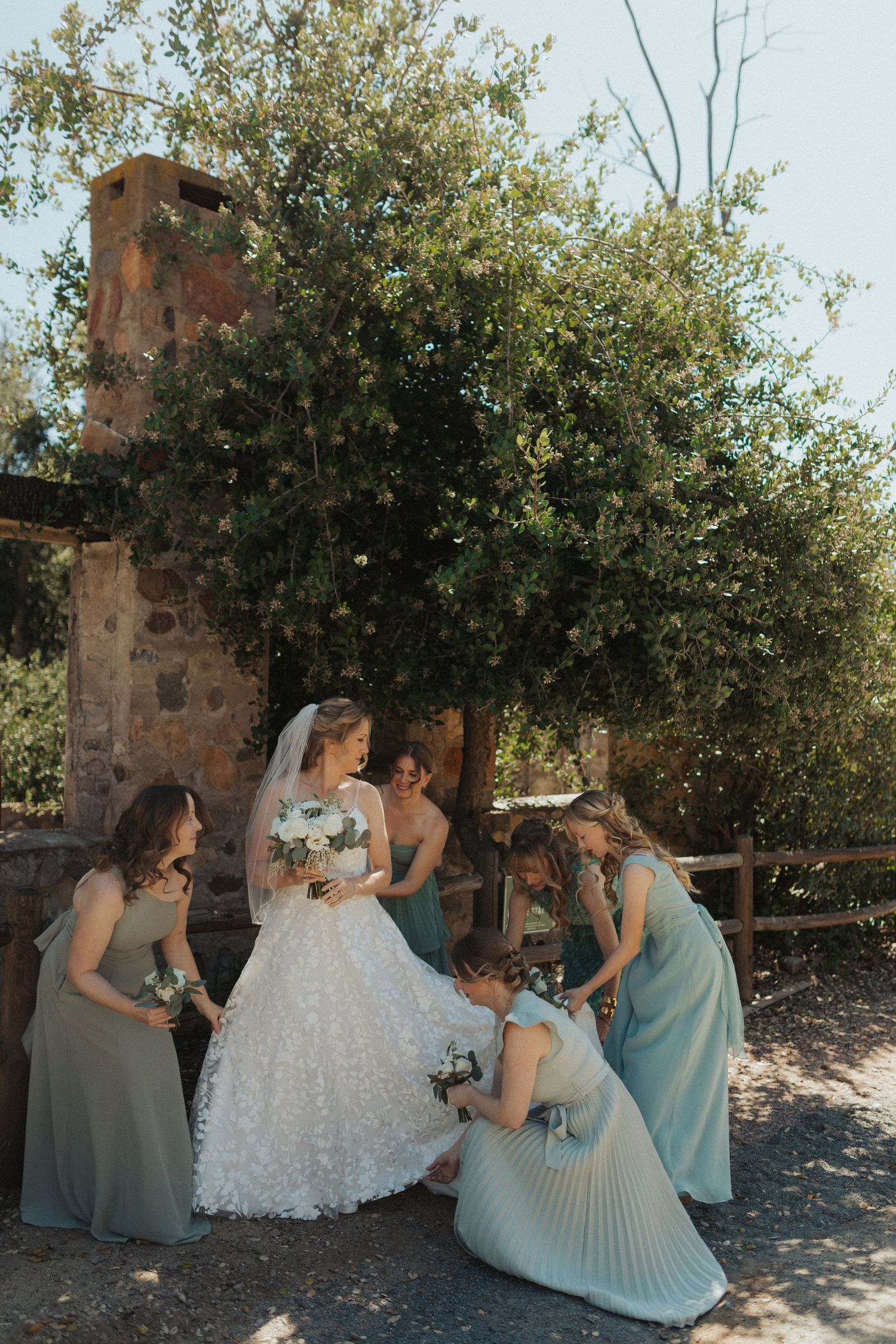 A bride in a floral wedding gown stands with her bridesmaids in sage green dresses near a stone outdoor setting.