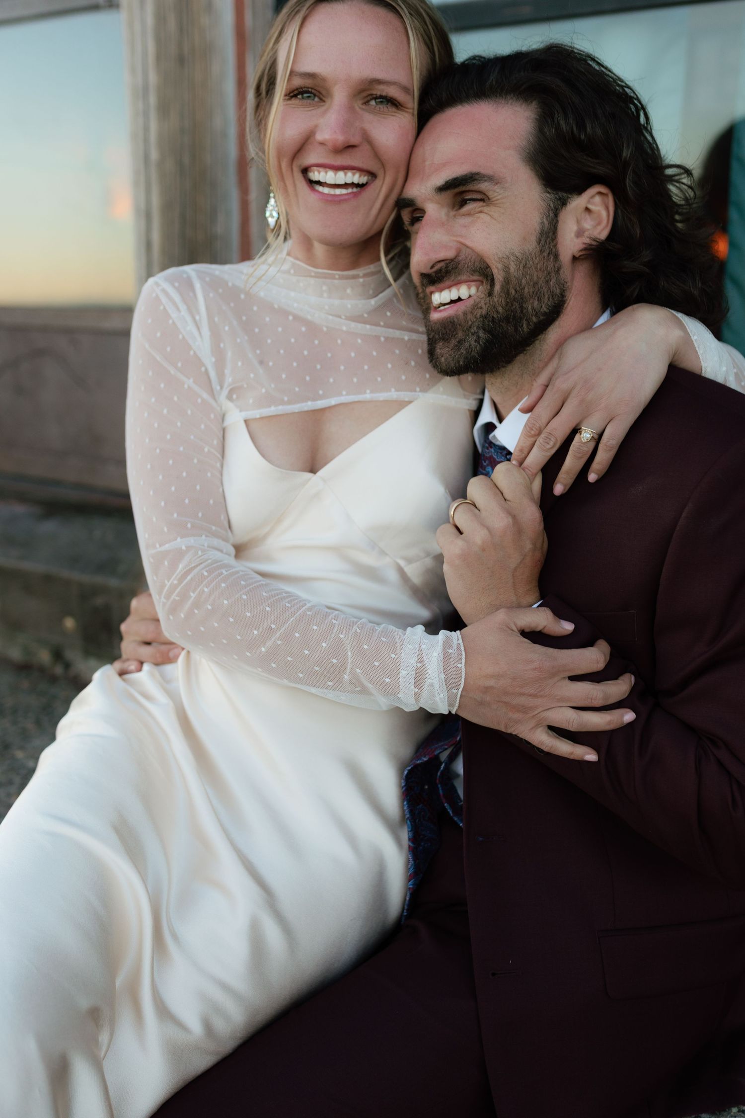 Laughing bride sits on grooms lap in a white gown and burgundy suit at sunset.