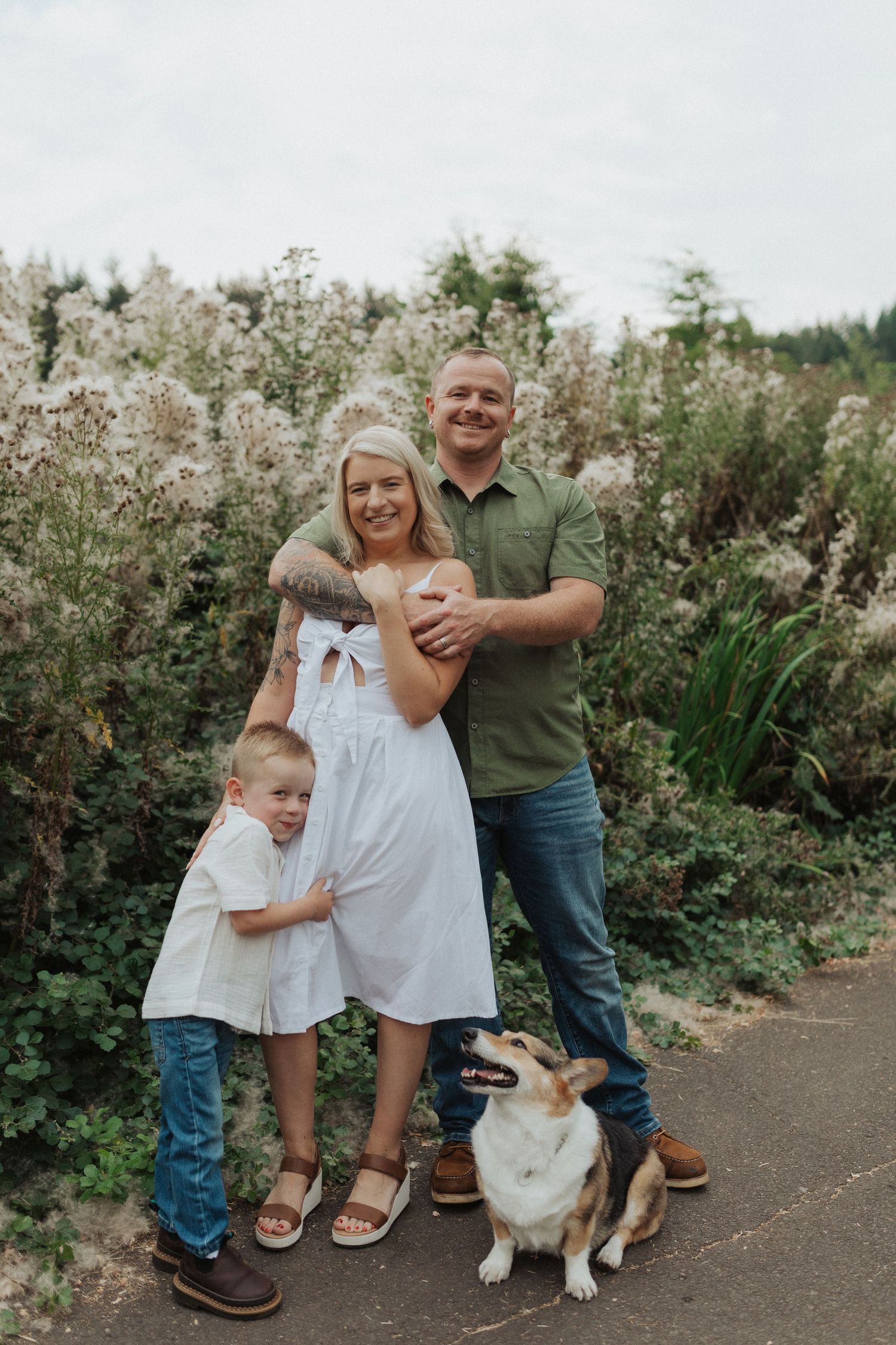 Happy family with young son and corgi dog posing in front of wildflower bushes outdoors.