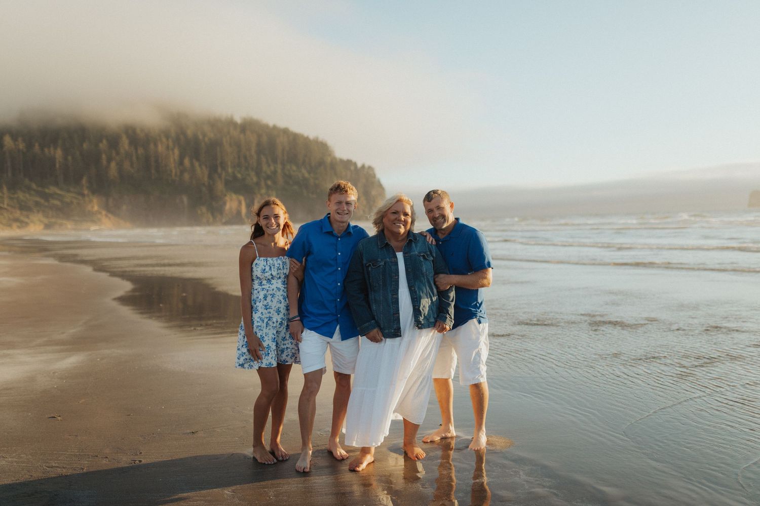 Happy family of four posing together on a sunny beach with misty forested cliffs behind them.