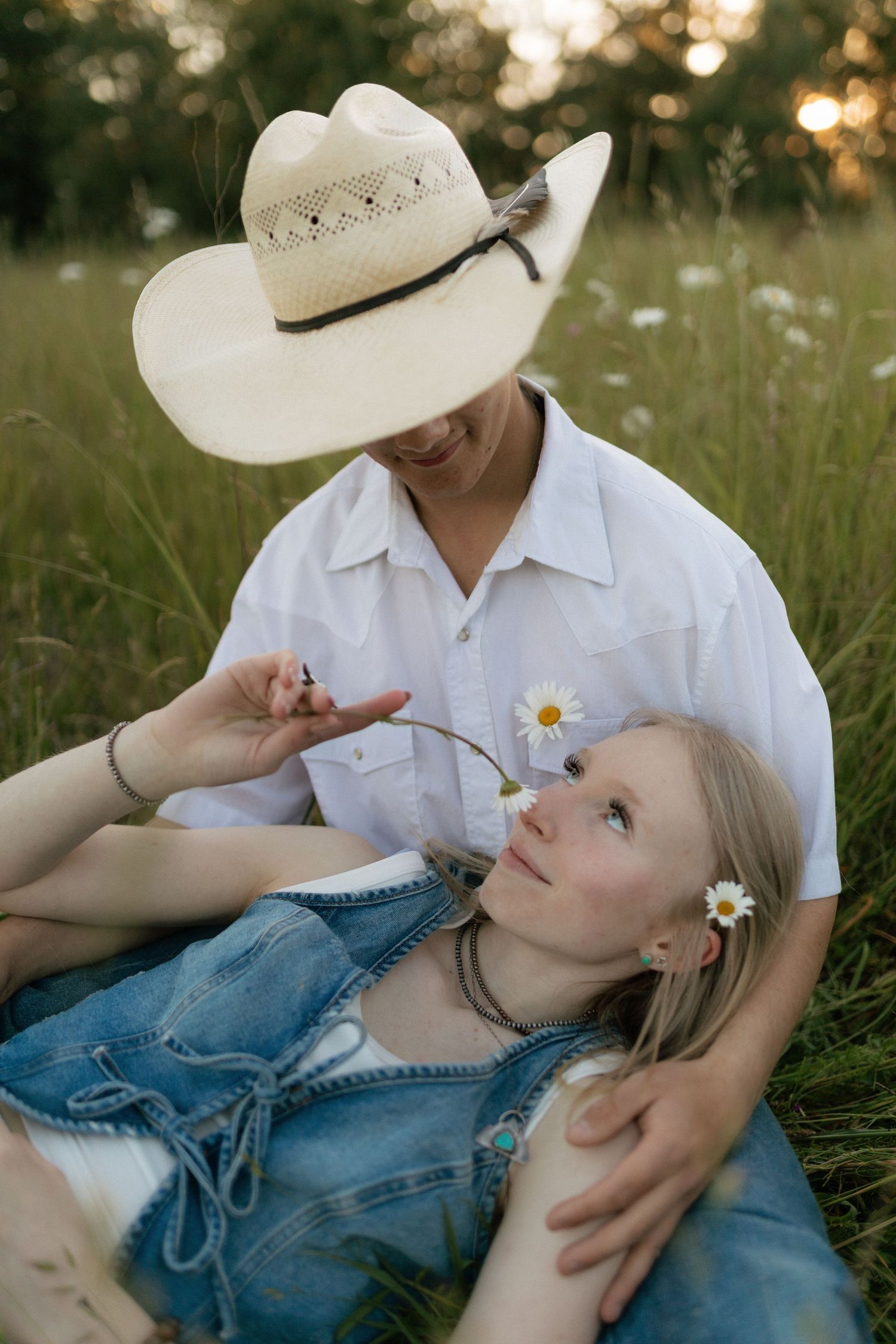 Man in cowboy hat holding daisy over smiling woman lying in his lap in a sunny meadow.
