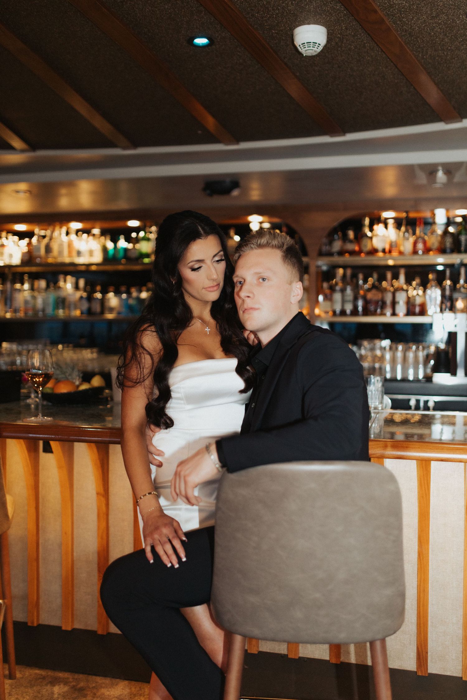 Elegant couple posing at a upscale bar, woman in white dress and man in black suit sitting on barstool.