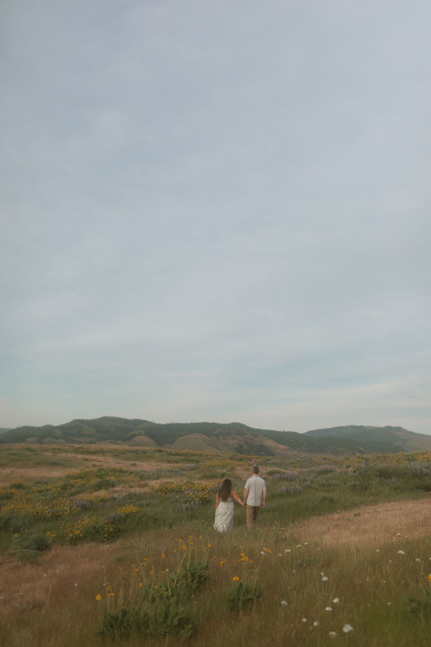 Couple walking hand in hand through a wildflower meadow with green hills under a cloudy sky.