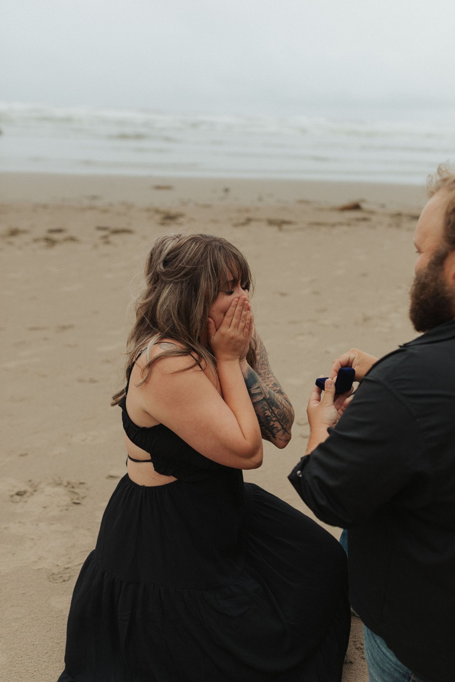 Emotional woman covering her face during beach marriage proposal, wearing black dress.