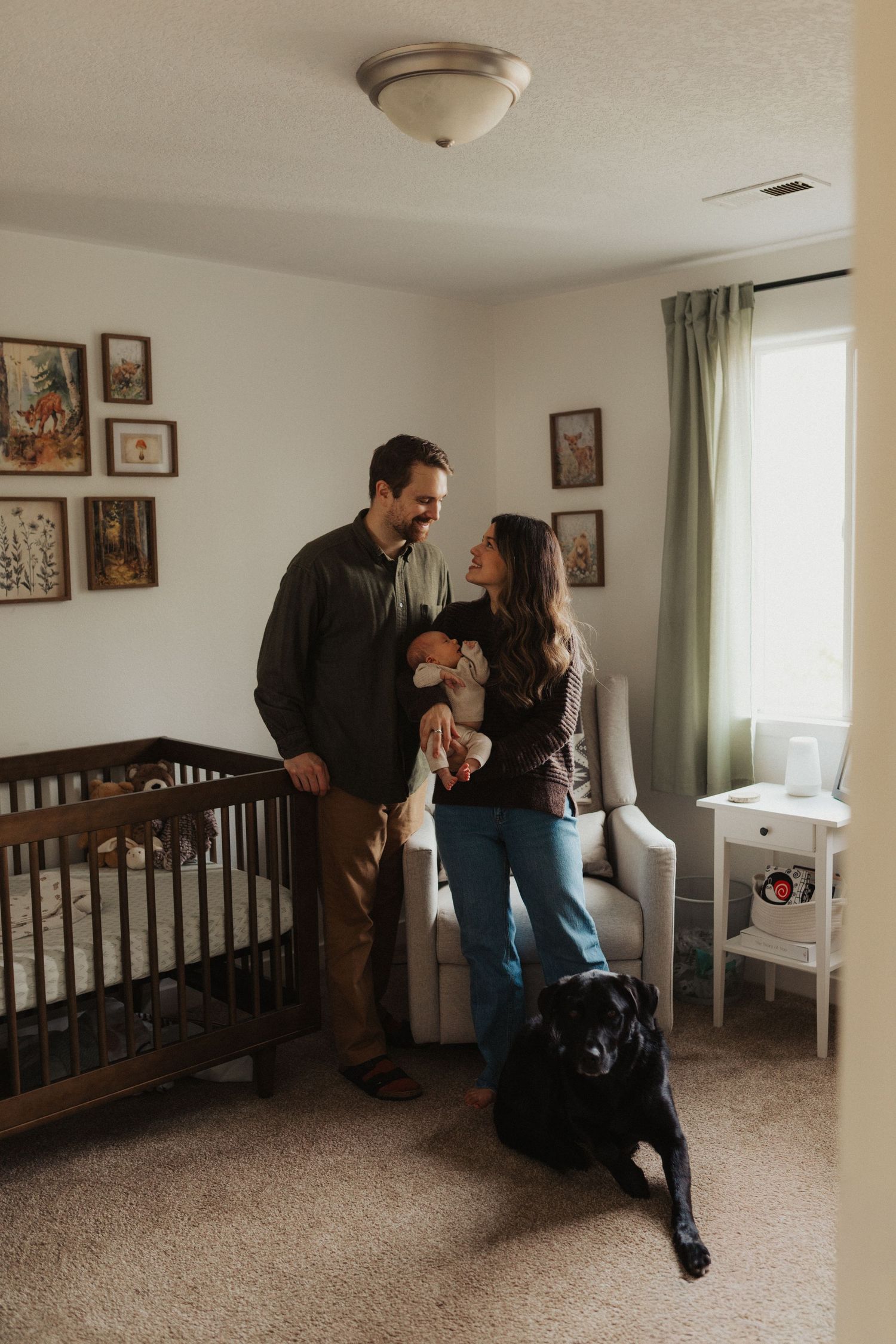 Young family with newborn baby and black Labrador dog standing in a decorated nursery room with a wooden crib.