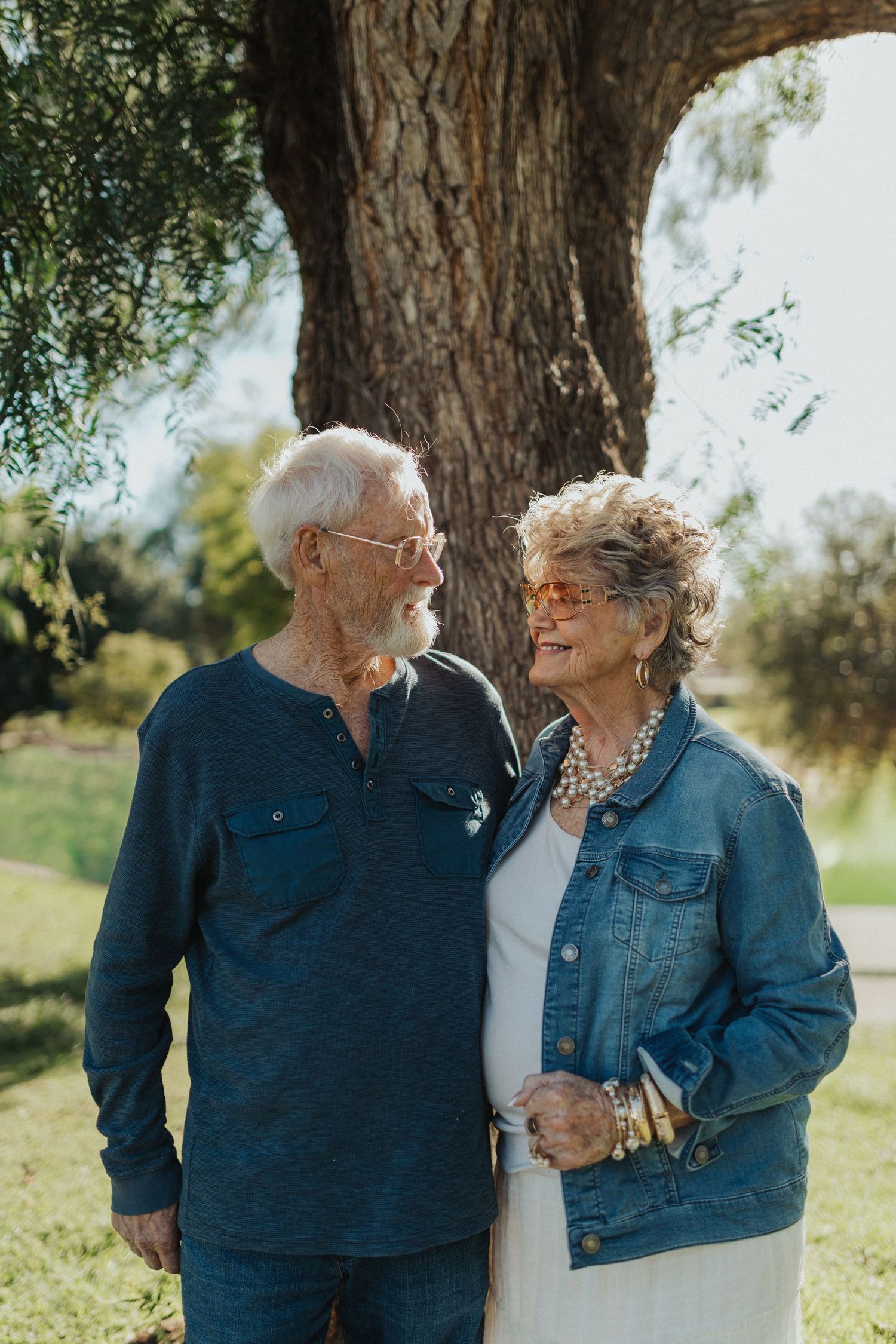 An elderly couple shares a loving glance while standing together near a large tree outdoors.