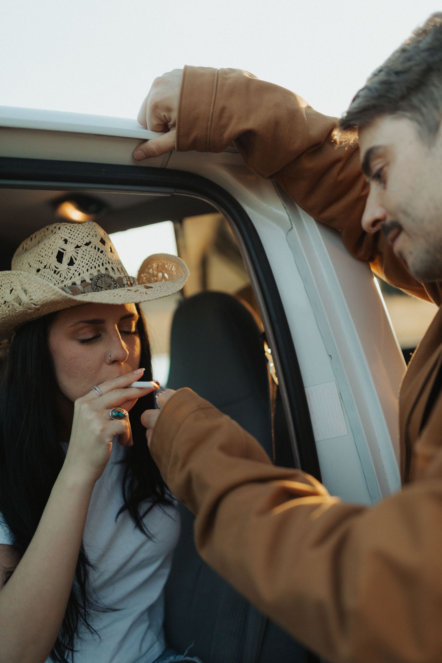 Man in brown jacket lighting a cigarette for woman in cowboy hat sitting in vintage pickup truck.