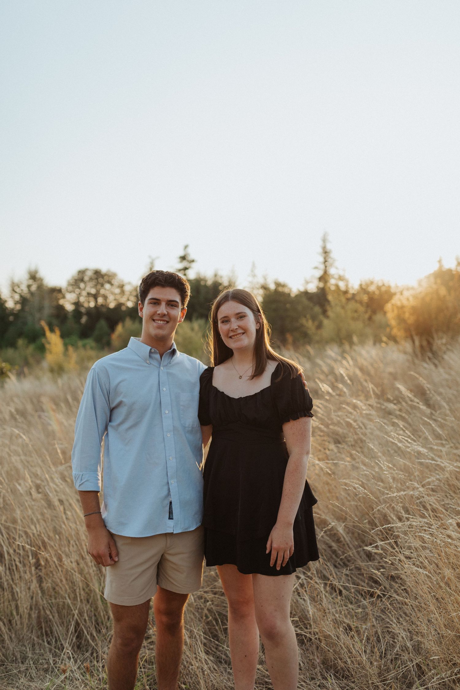 Young man and woman smiling together in a sunlit golden grass field at sunset.