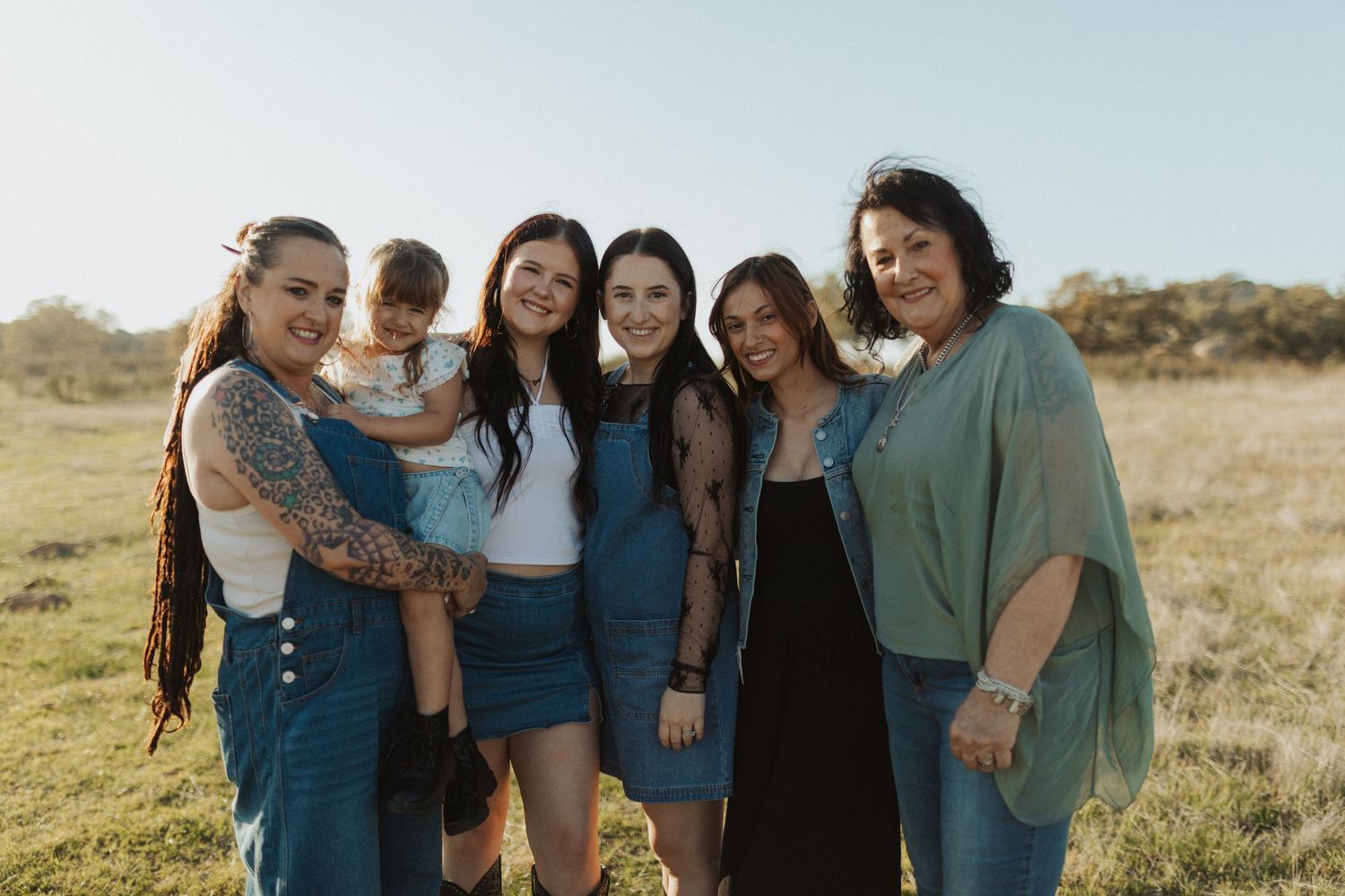 Group of five women and a toddler smiling together in a sunny open field outdoors.