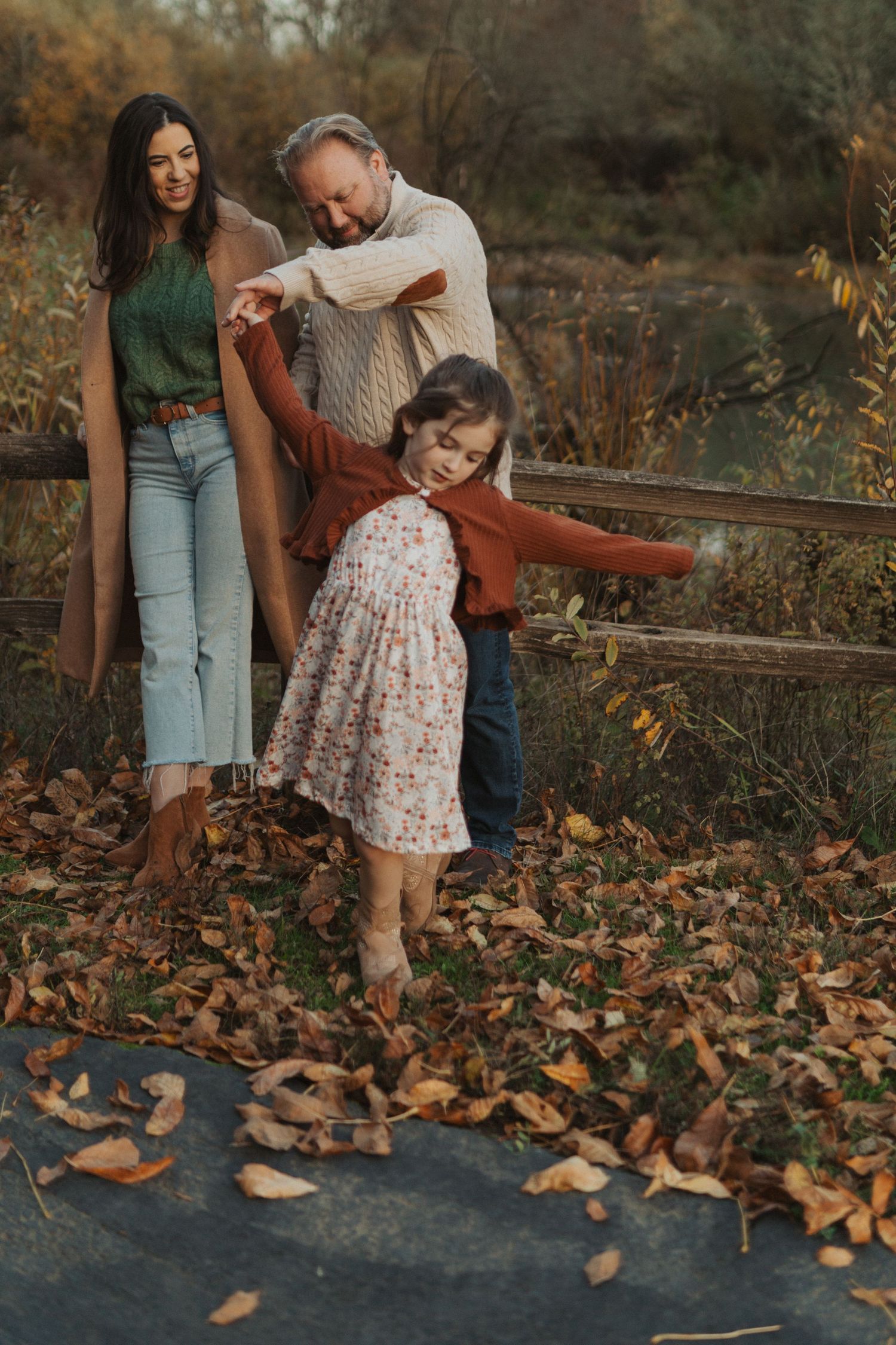 Parents help their young daughter balance on a surface covered in fallen autumn leaves outdoors.