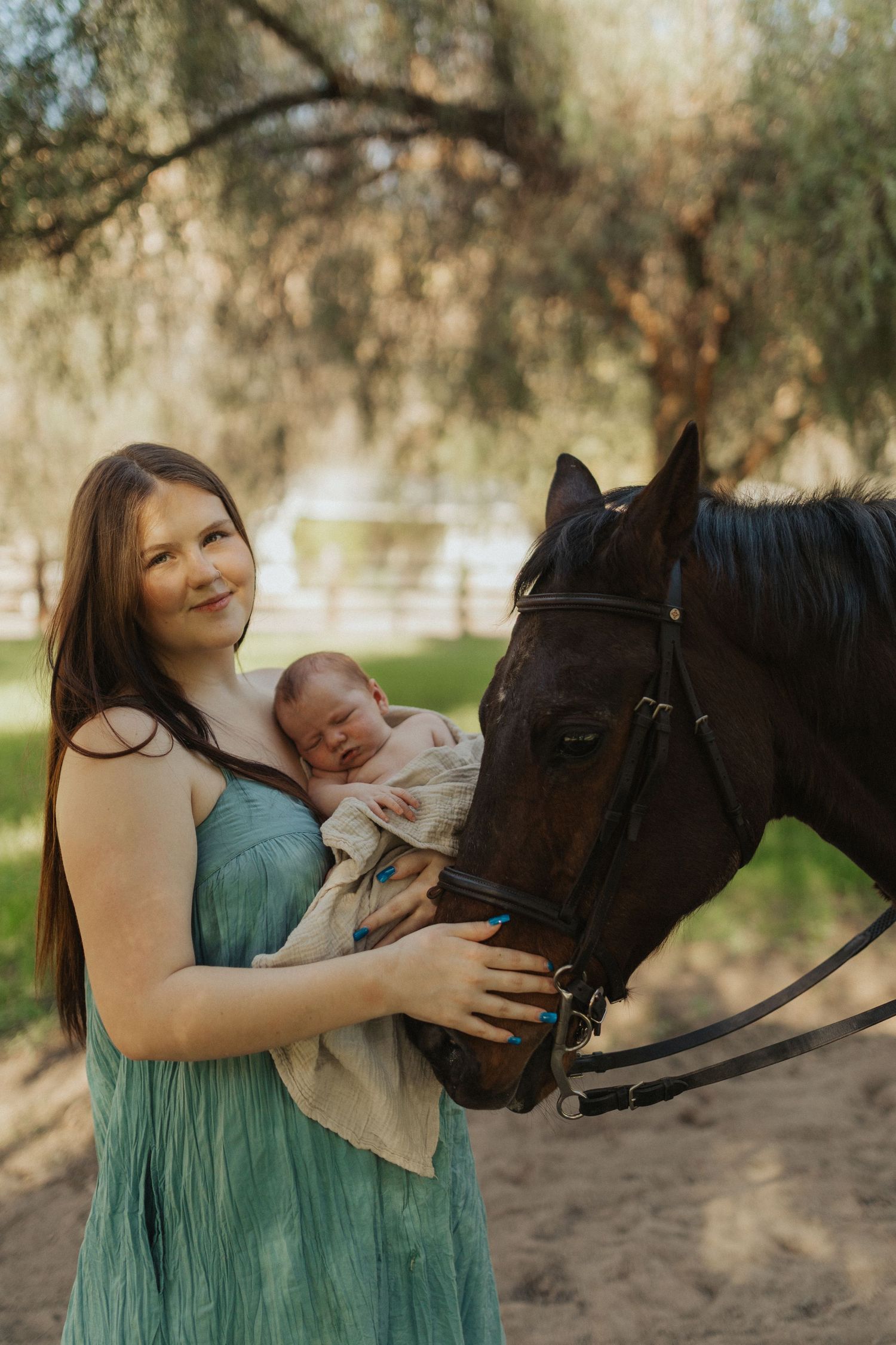 Smiling mother holding newborn baby while dark horse nuzzles them in sunny outdoor setting.