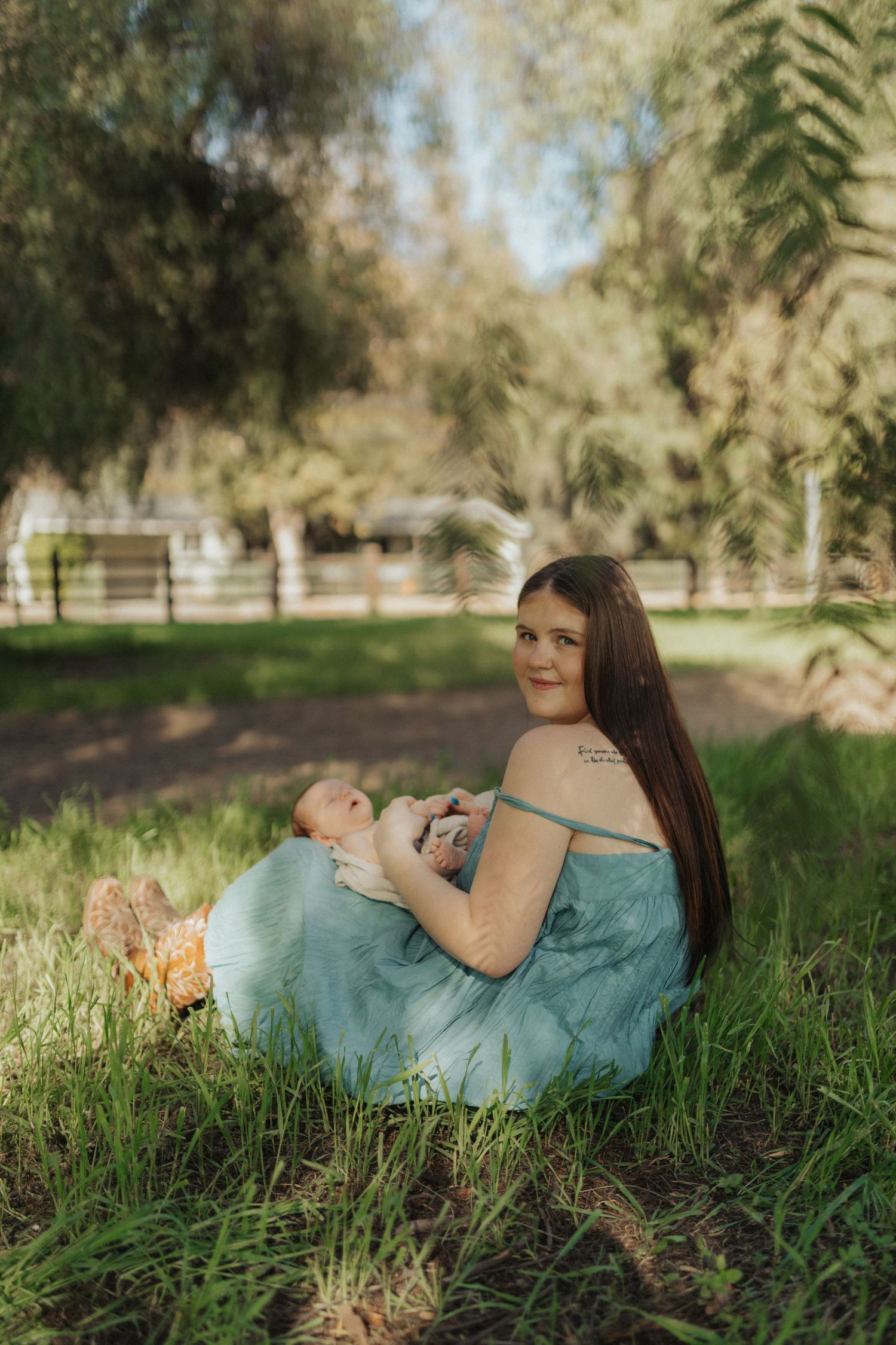 Smiling mother in teal dress sitting on lush green grass holding newborn baby in sunny park.