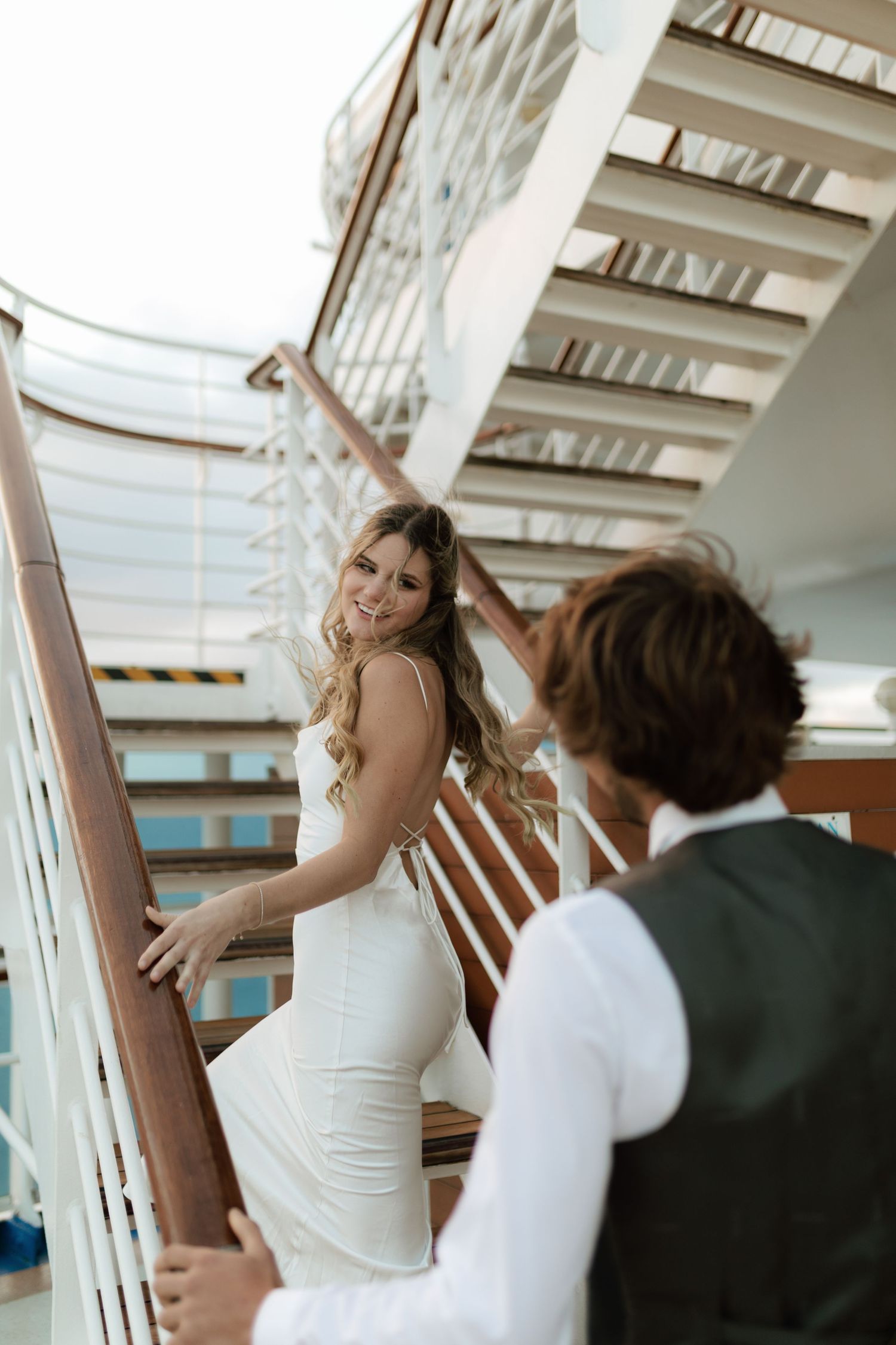 Smiling bride in a white gown looking back at groom while ascending cruise ship stairs.