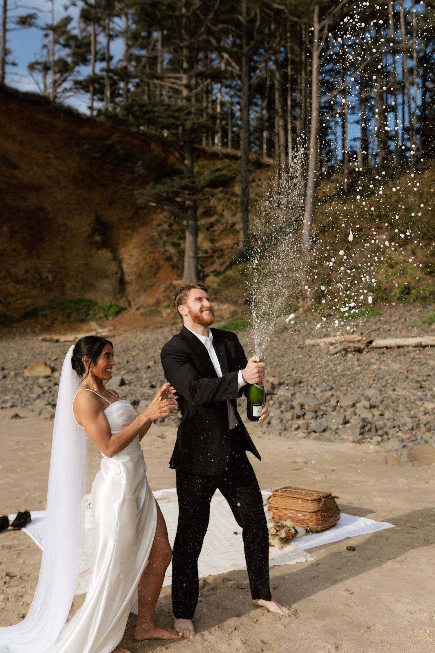 Groom pops champagne bottle as bride laughs beside him on beach surrounded by trees and picnic setup.