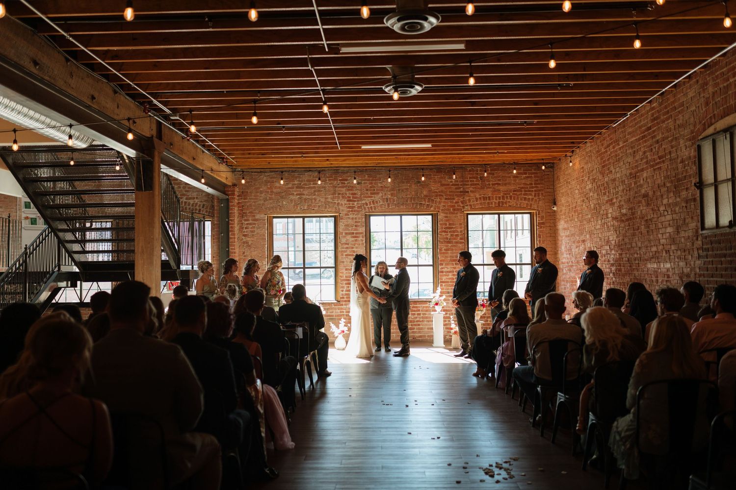 Wide angle view of intimate wedding ceremony in industrial brick venue with string lights overhead.
