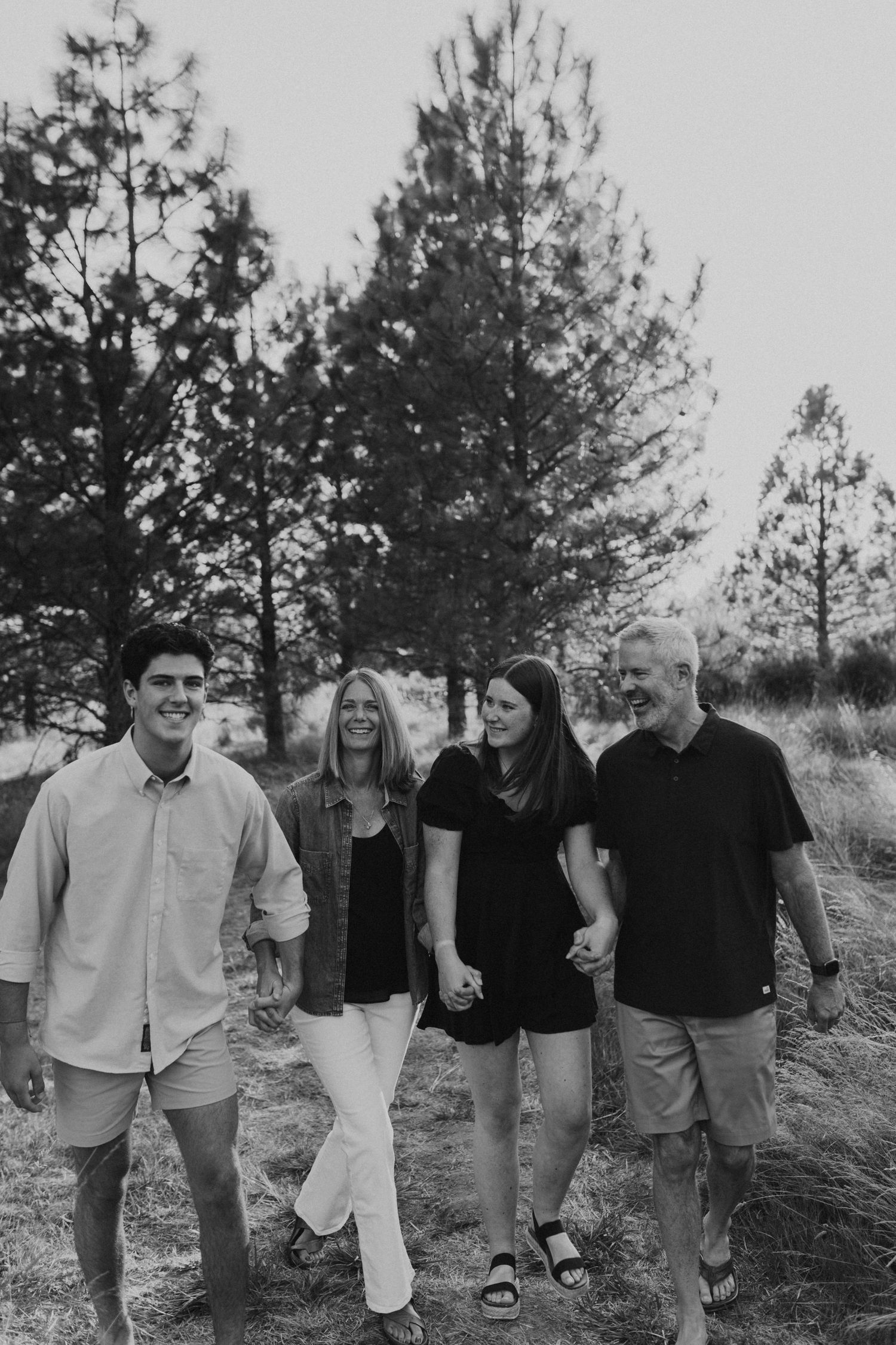 Black and white photo of a happy family of four walking outdoors near pine trees.