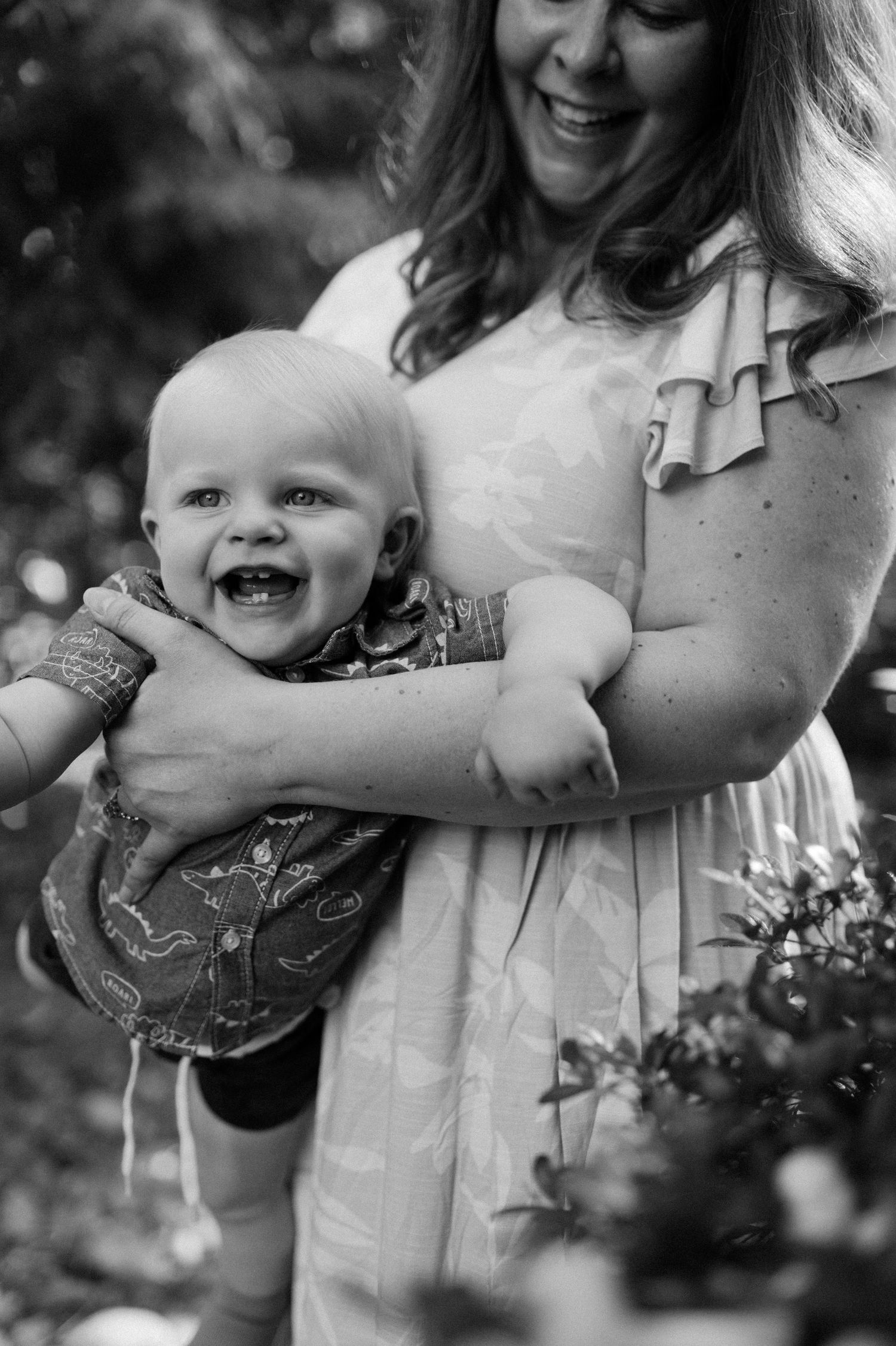 Black and white portrait of laughing baby being held by smiling mother in outdoor garden setting.