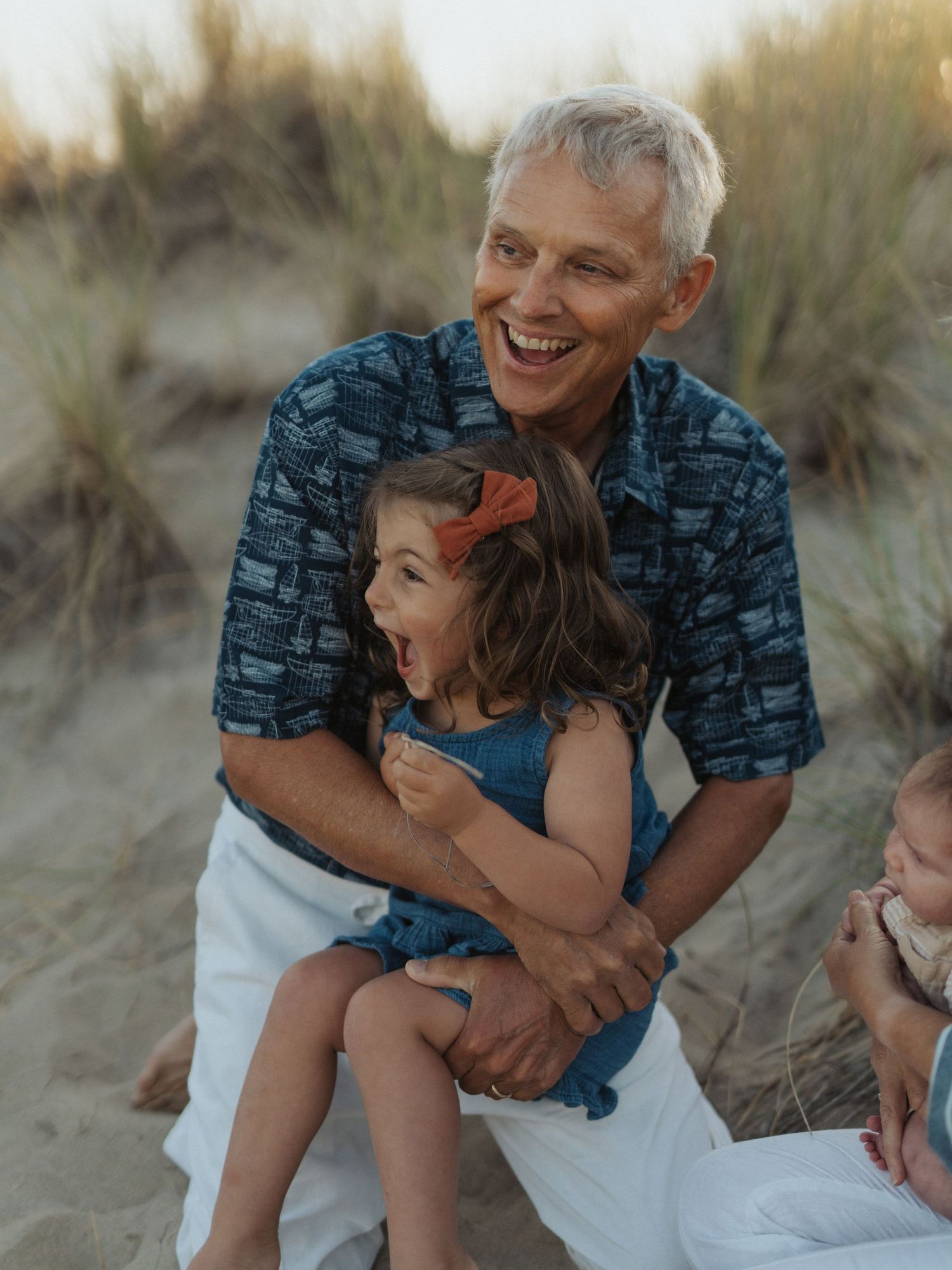 Laughing grandfather in blue patterned shirt holds giggling young granddaughter on sandy beach.