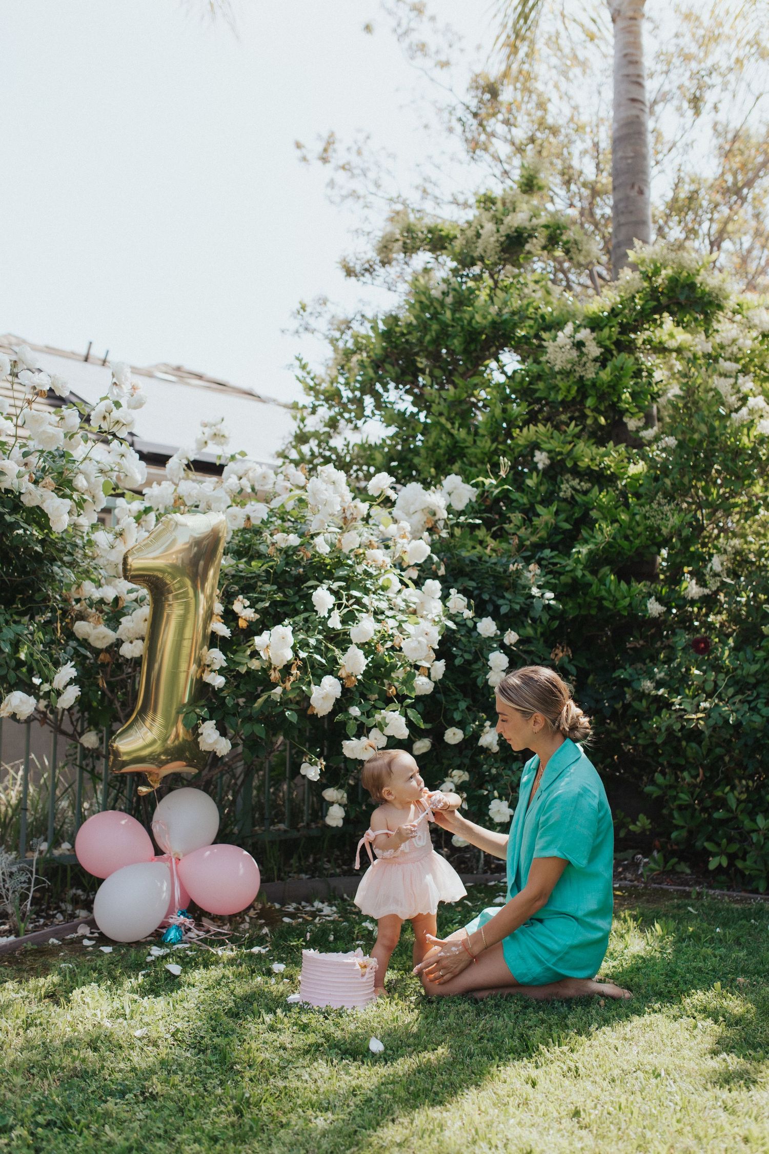 Mother in teal dress sits on grass helping toddler stand near number one balloon in garden.