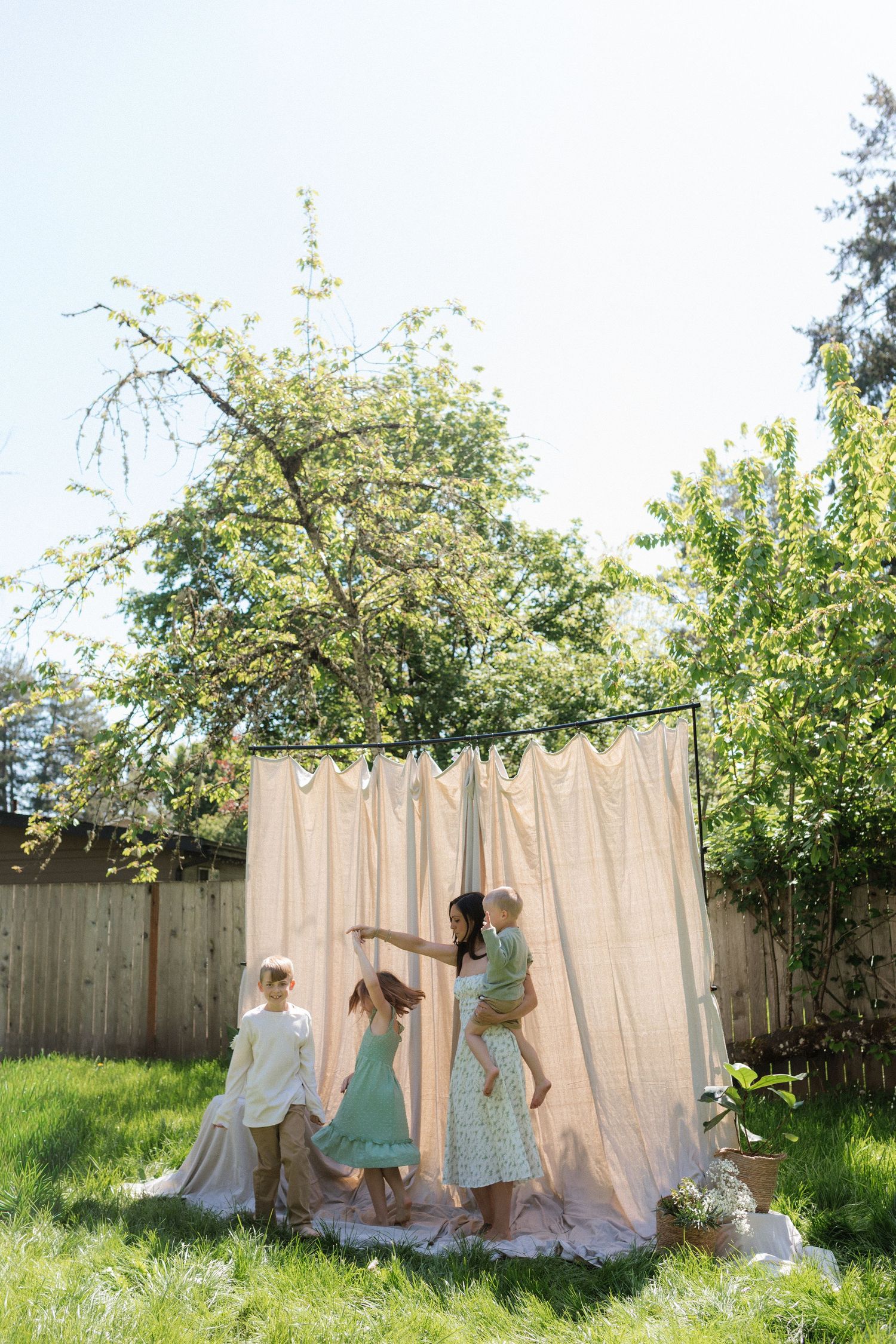 A mother dances playfully with her children in a sunny backyard near a draped fabric backdrop.