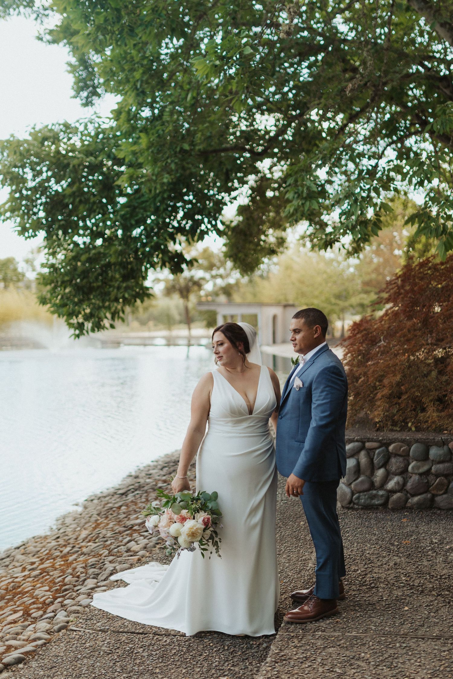 Bride in white gown holding bouquet stands with groom in blue suit by a serene lakeside under lush green trees.