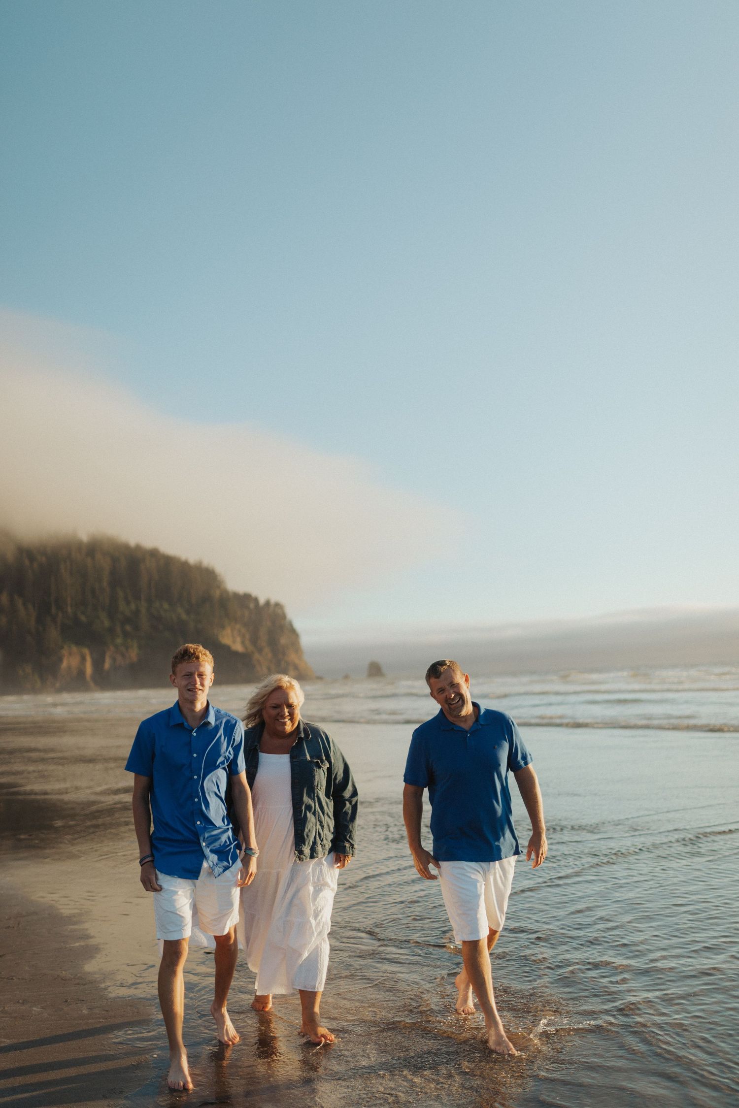 Three family members walking barefoot through shallow ocean water on a misty Pacific coast beach.